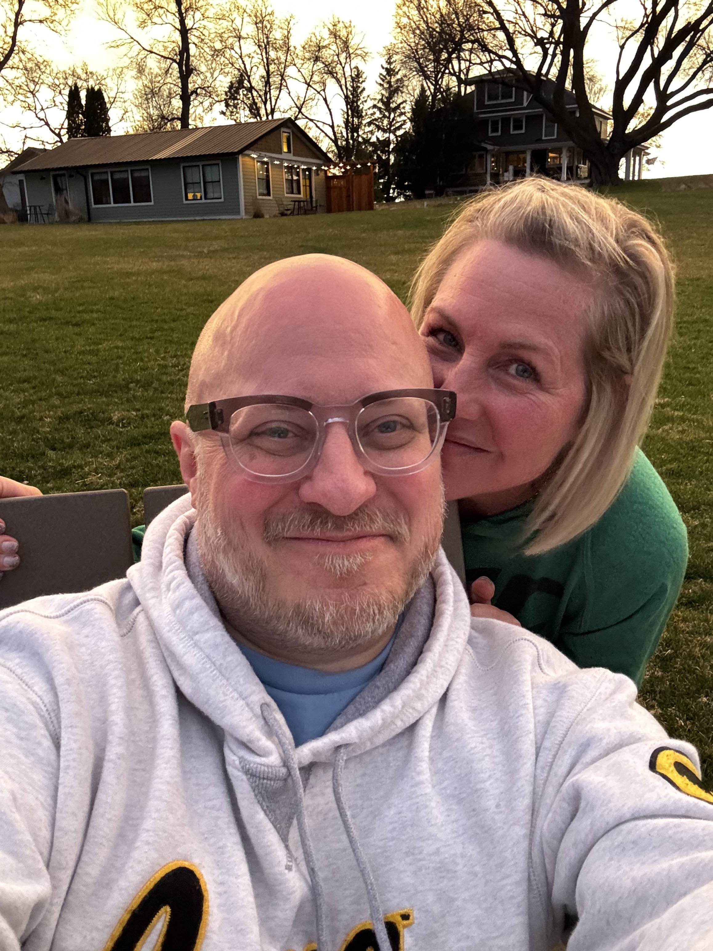 A man and woman taking a selfie outdoors on a grassy hill with trees and houses in the background during sunset.