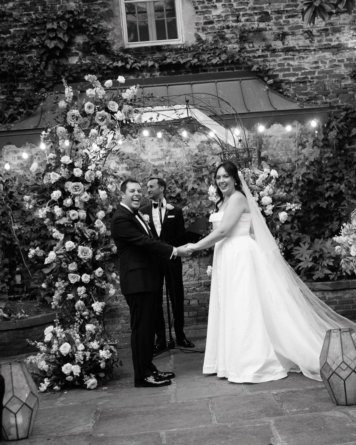 Bride and groom laughing during their courtyard ceremony at Brennan’s Restaurant in New Orleans, standing beneath a floral arch surrounded by ivy and string lights.