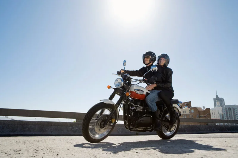 Two people riding a motorcycle, with a city in the background.