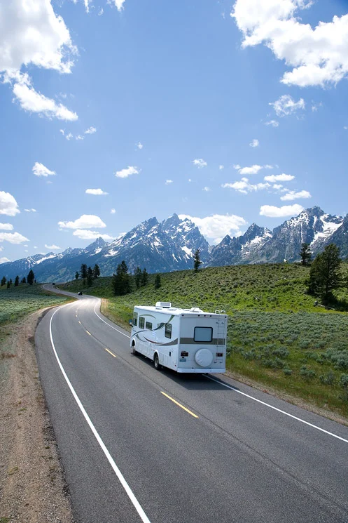 Recreational Vehicle driving into the distance, with mountains and forest in the background.