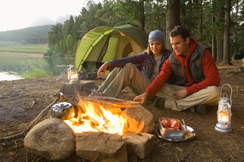 A man and a woman camping; cooking over a fire in the foreground, with a tent and gear behind them.