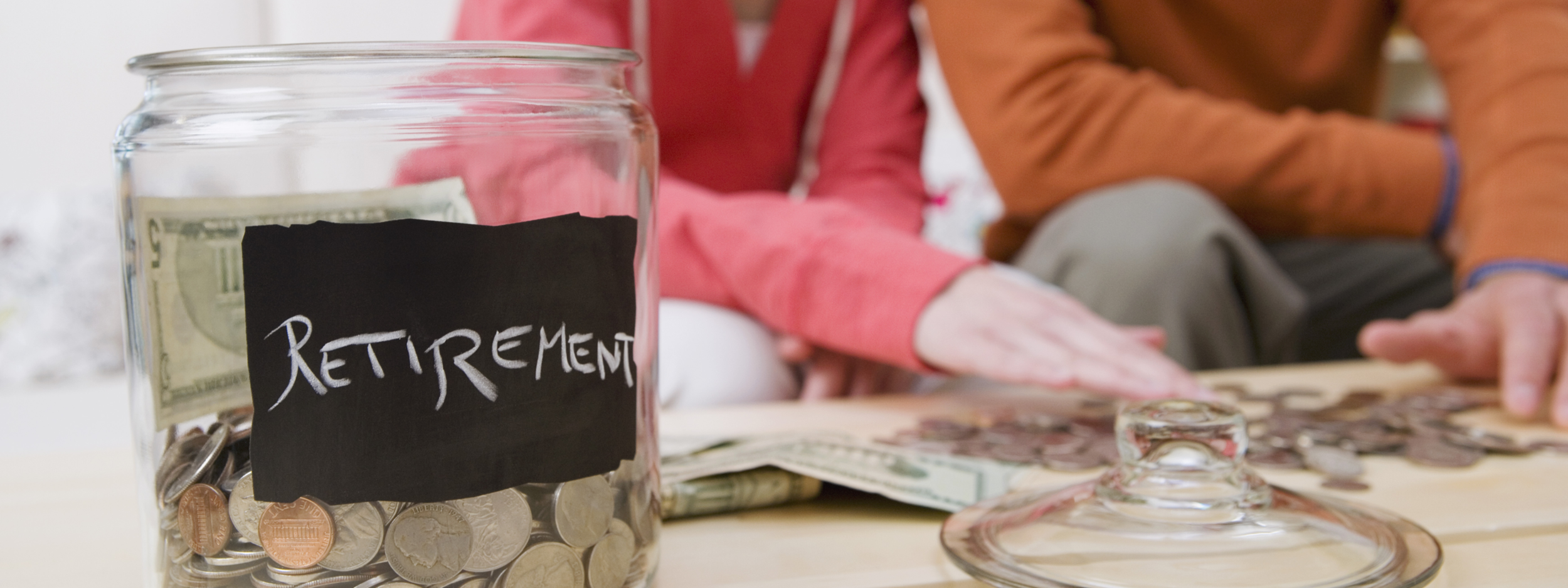 A jar labeled “retirement” with dollars and coins in it, with two people counting money in the background