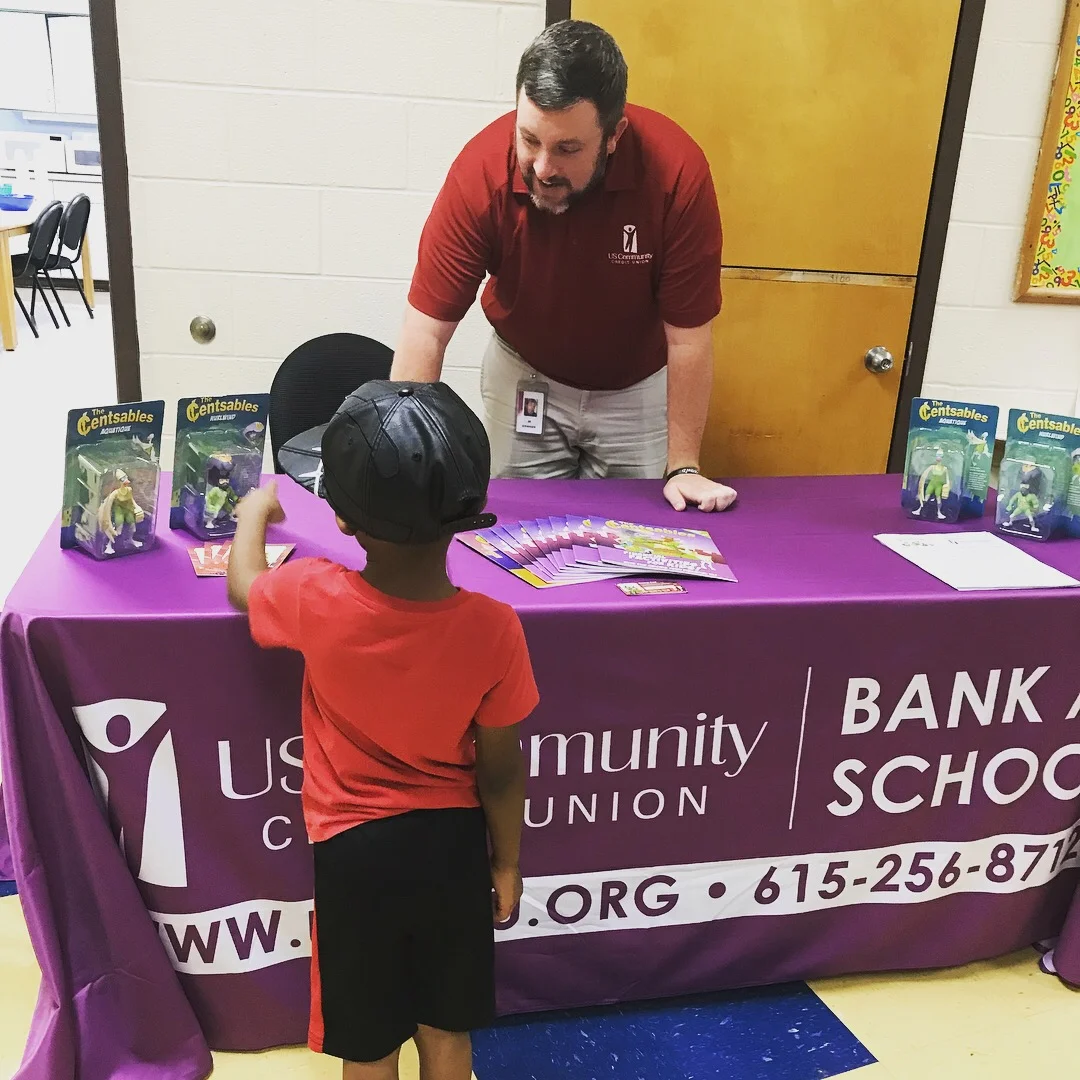 A elementary school student pointing at a Censtables action figure at a USCCU Bank to School table