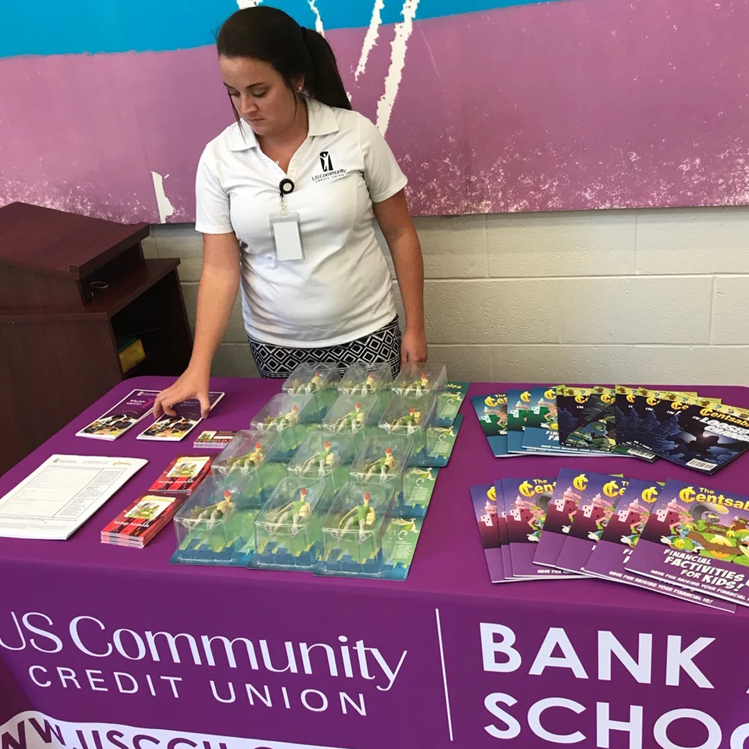 A USCCU employee arranging Censtables materials on a table with a sign reading “US Community Credit Union Bank to School”