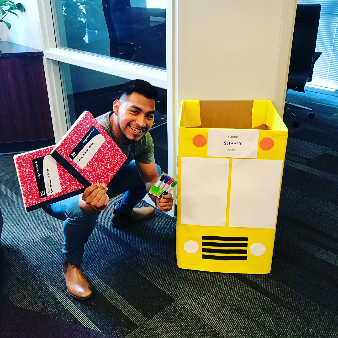 A smiling man holding pens and notebooks in front of a school donation bin.
