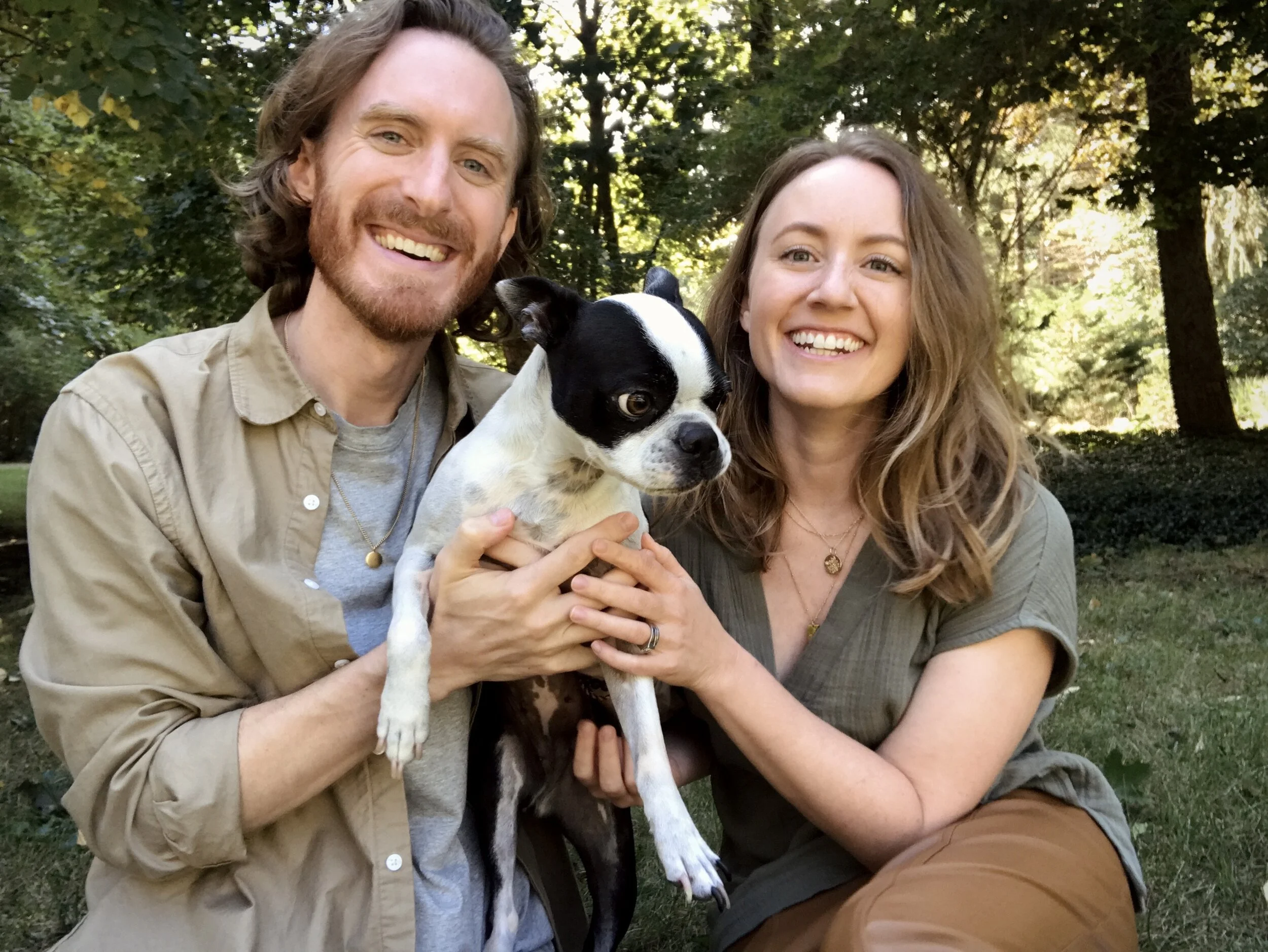 Somatic therapists Anne and Steve Truppe outdoors holding a Boston Terrier dog with a black and white coat, surrounded by trees and sunlight.