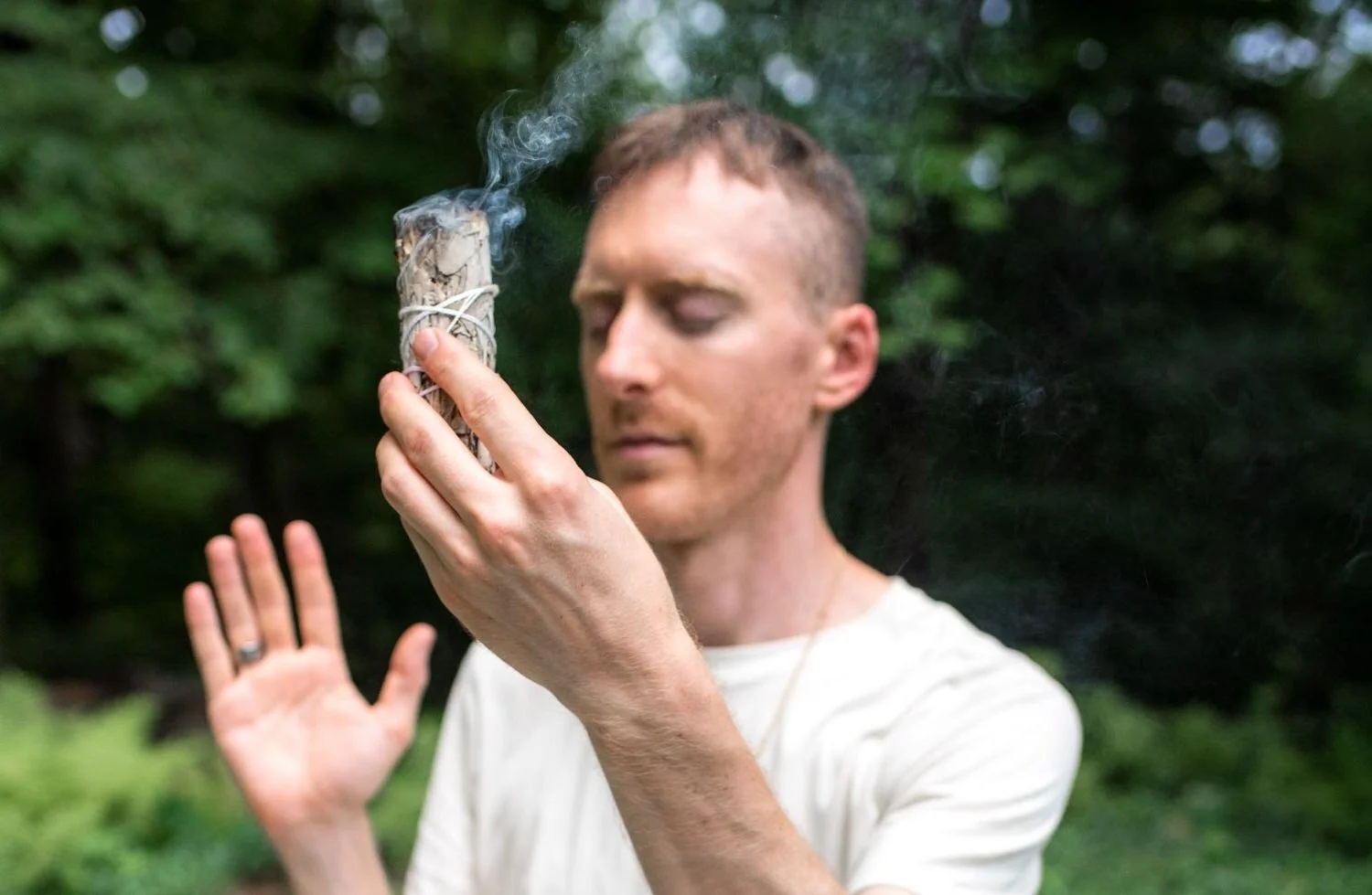 A man in a white shirt holds a lit smudge stick emitting smoke, with his eyes closed and his other hand raised in a prayer-like gesture, outdoors with green trees in the background.
