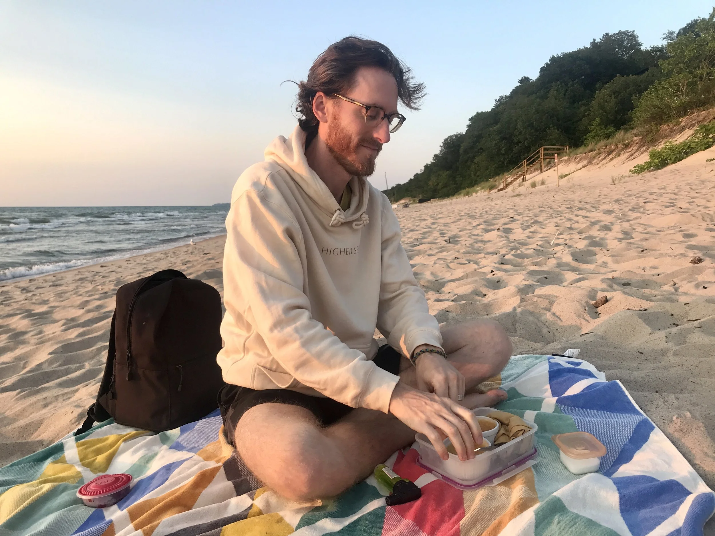 A man with glasses and a beard wearing a beige hoodie and shorts, sitting cross-legged on a colorful towel on the sandy beach during sunset, packing a lunchbox with food.