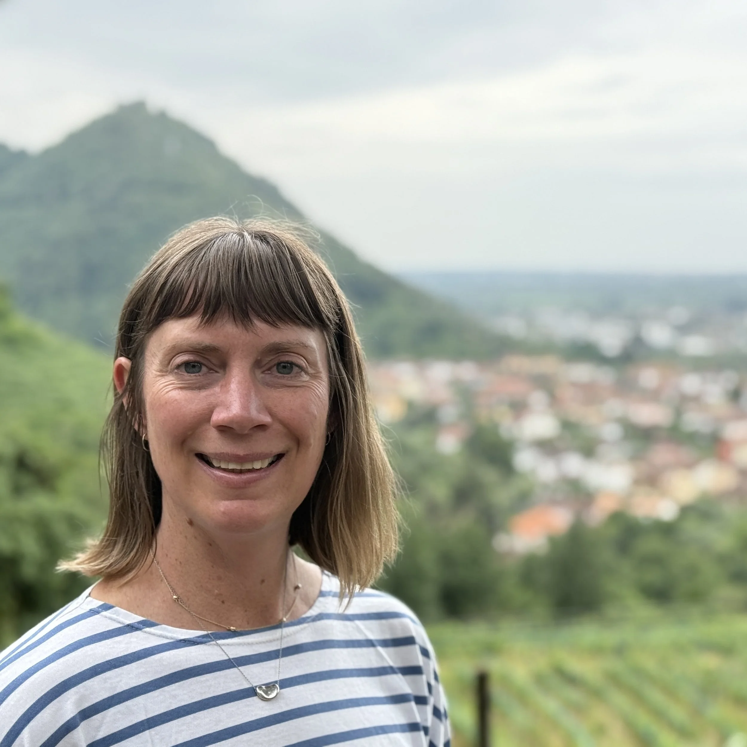 Sarah Kotcher. A woman with shoulder-length brown hair and wearing a blue and white striped shirt, smiling outdoors with mountains and a small town in the background.
