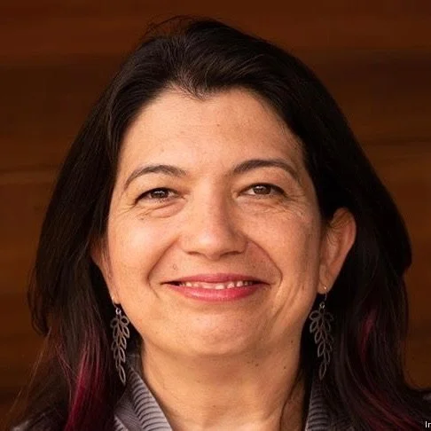 Olgica Bakajin. A middle-aged woman with shoulder-length dark hair, smiling, wearing earrings and a light-colored top, against a wooden background.