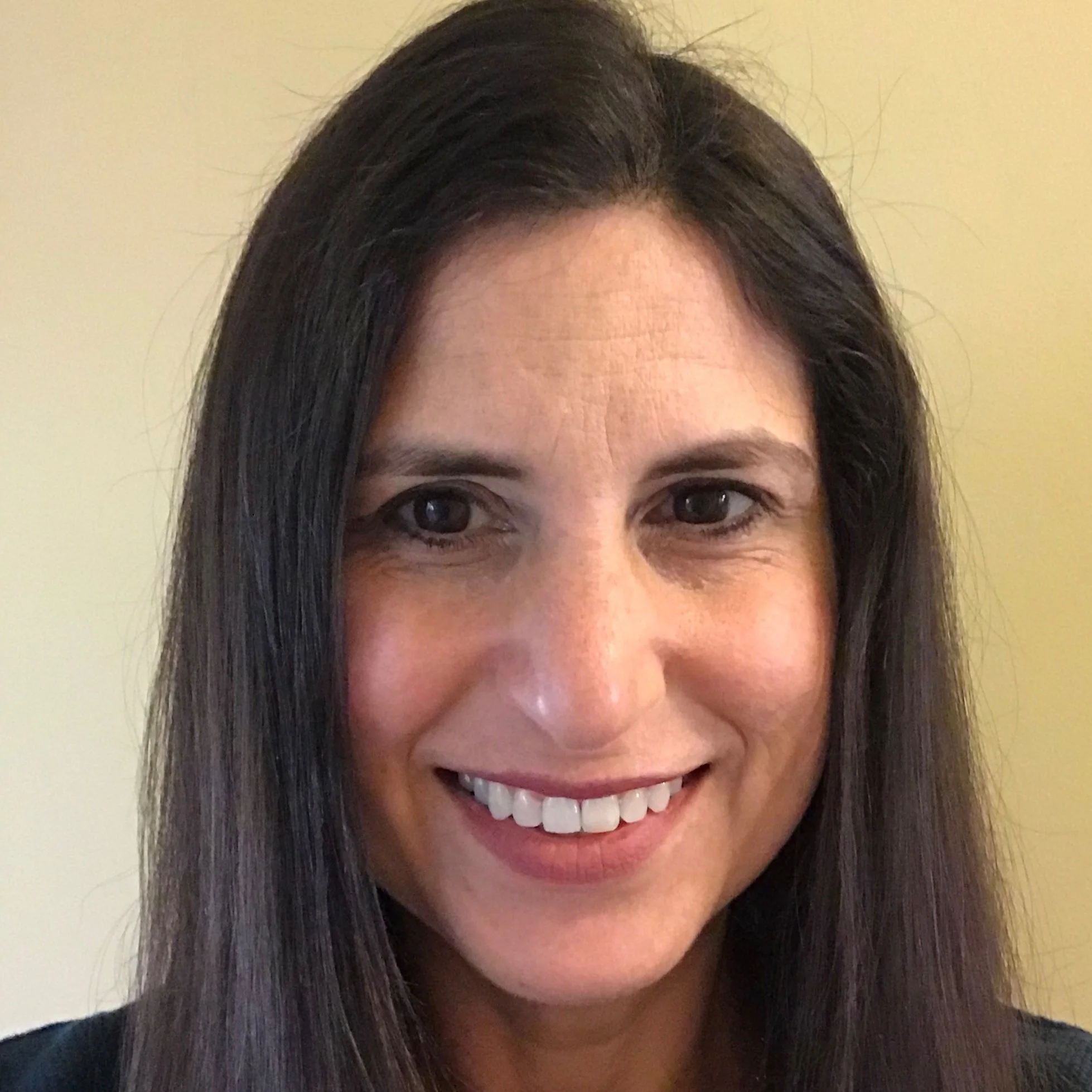 Michele Ramos Walker. Close-up portrait of a woman with long dark hair smiling, showing teeth, against a plain yellow background.