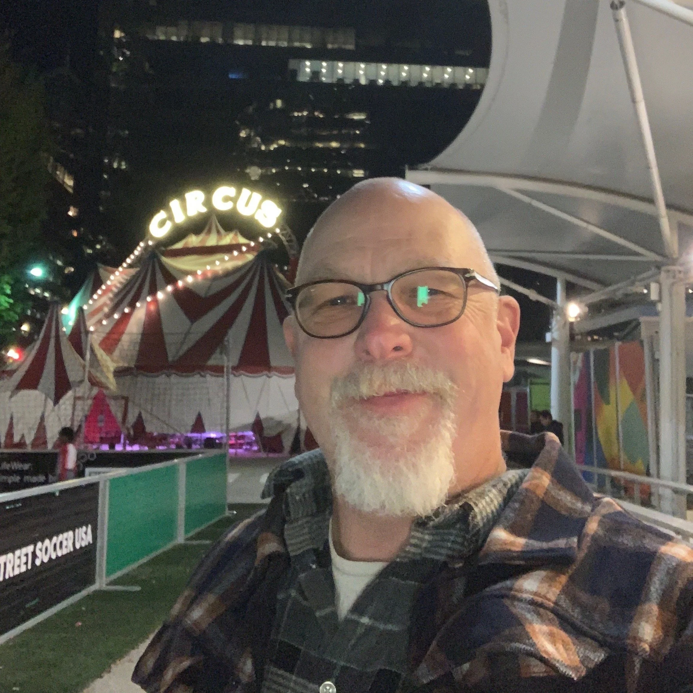 Eddie Scher. A man with glasses and a goatee taking a selfie at a circus-themed carnival at night, with a large striped tent and illuminated 'CIRCUS' sign in the background.