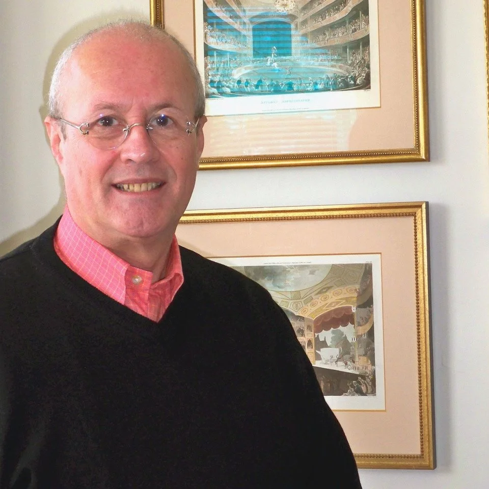 Dominque Jando. A man with glasses and a bald head, smiling, wearing a pink collared shirt and a black sweater, standing in front of framed artwork on a wall.