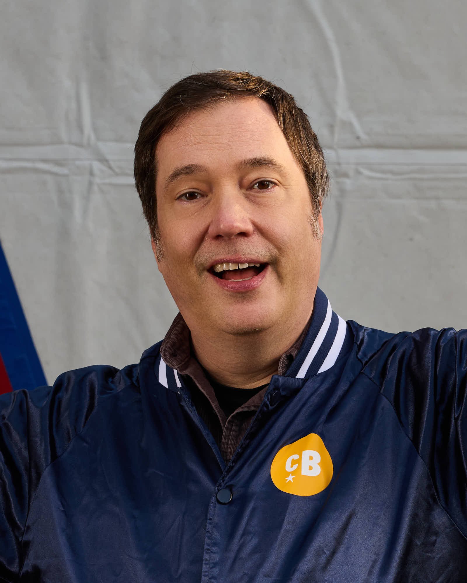 Trumpet player Ian Carey, wearing a navy blue jacket with a yellow sticker that has 'CB' written on it, holding a hockey stick over his shoulder, standing in front of a circus tent.