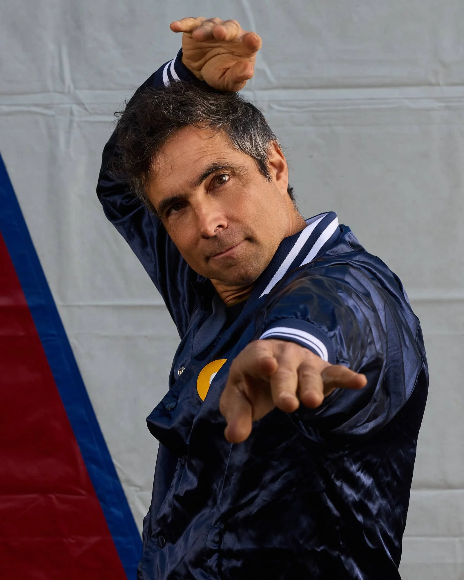 A man with dark curly hair and a slight smile, wearing a dark blue satin jacket with white stripes on the collar and the words 'Circus Bella' embroidered in gold on the chest, is standing in front of a Circus Tent.