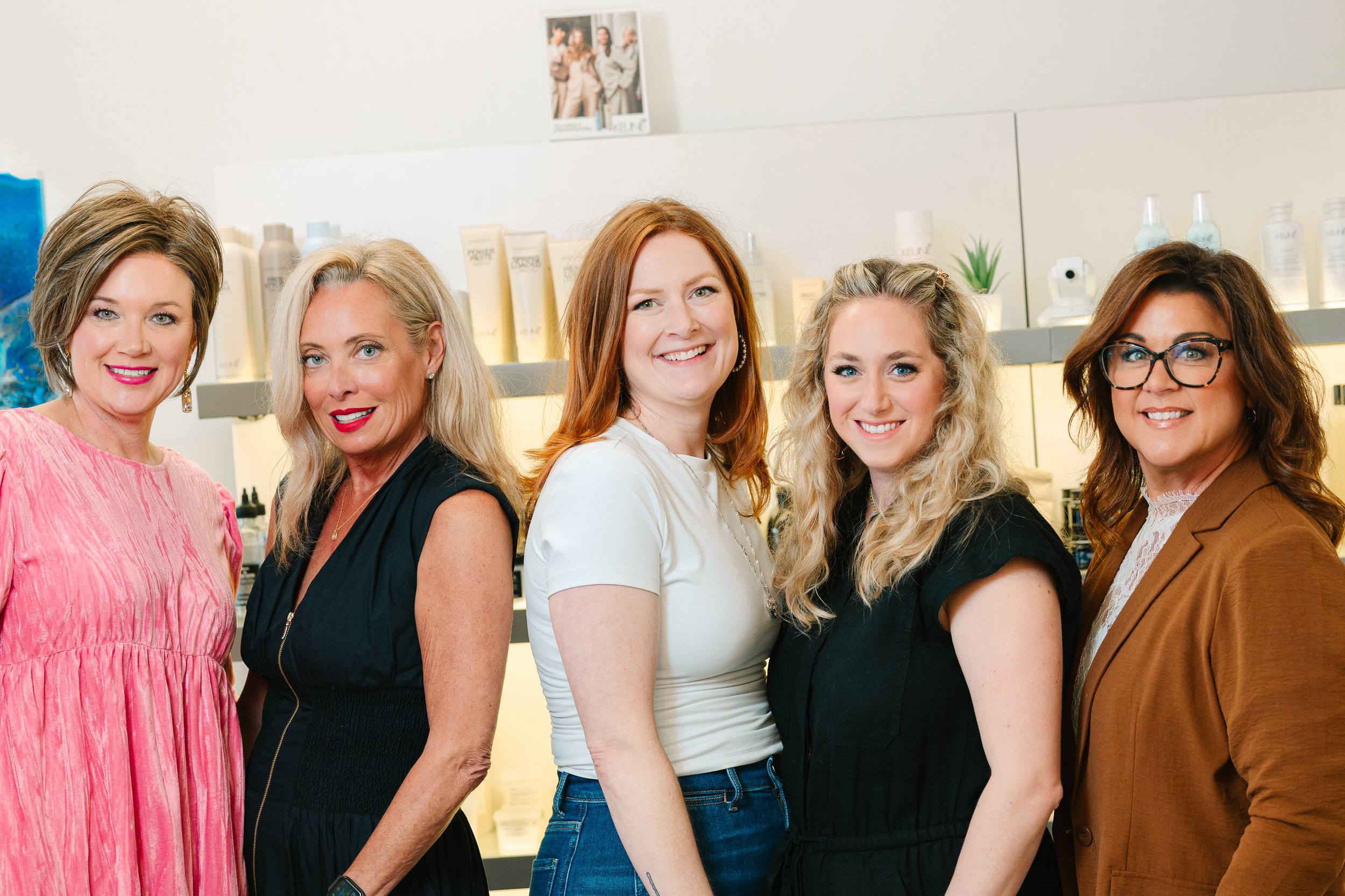 Five women standing together in a store, smiling at the camera, with shelves of hair products behind them.