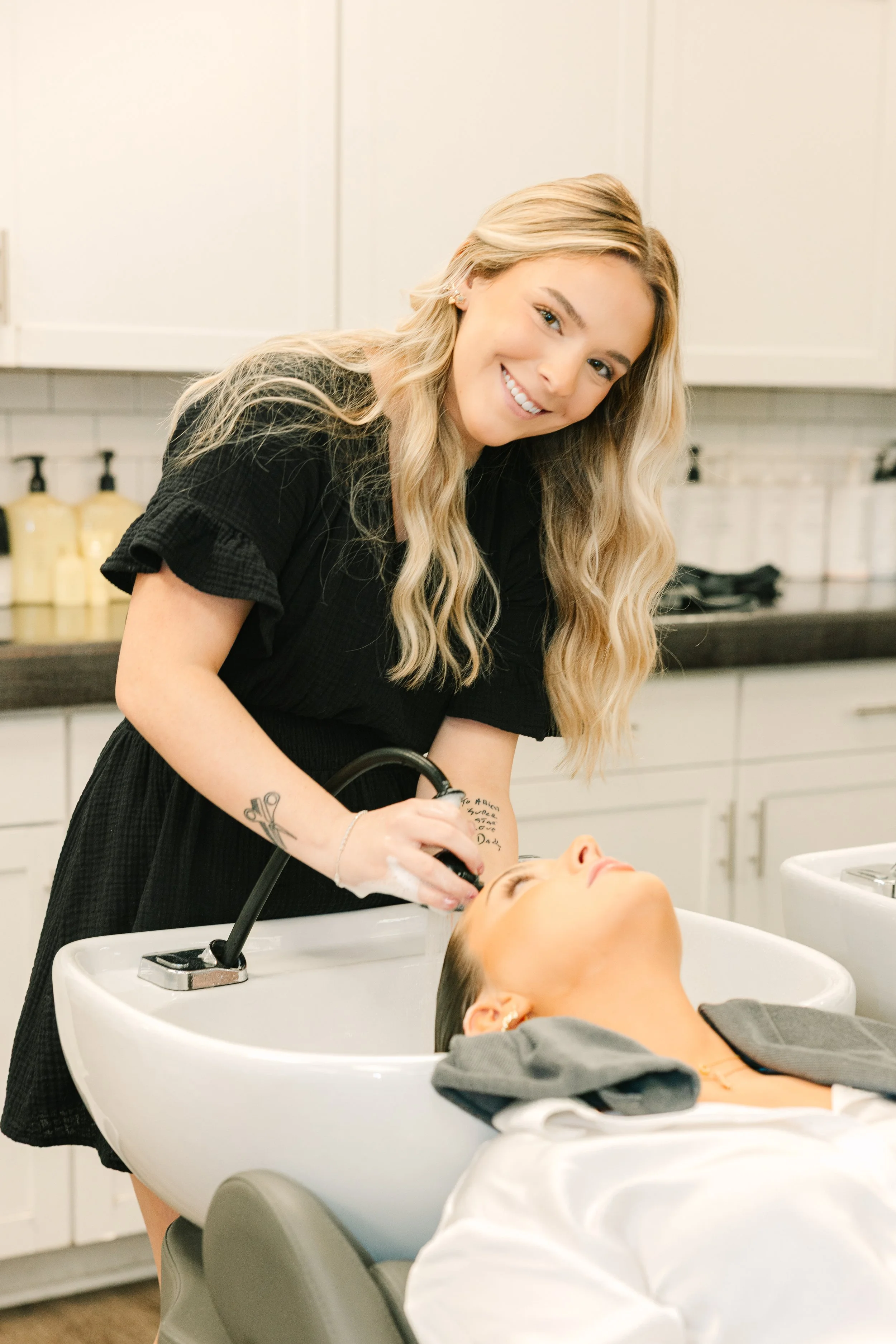 A woman with long blonde wavy hair washing a woman's hair in a salon sink.