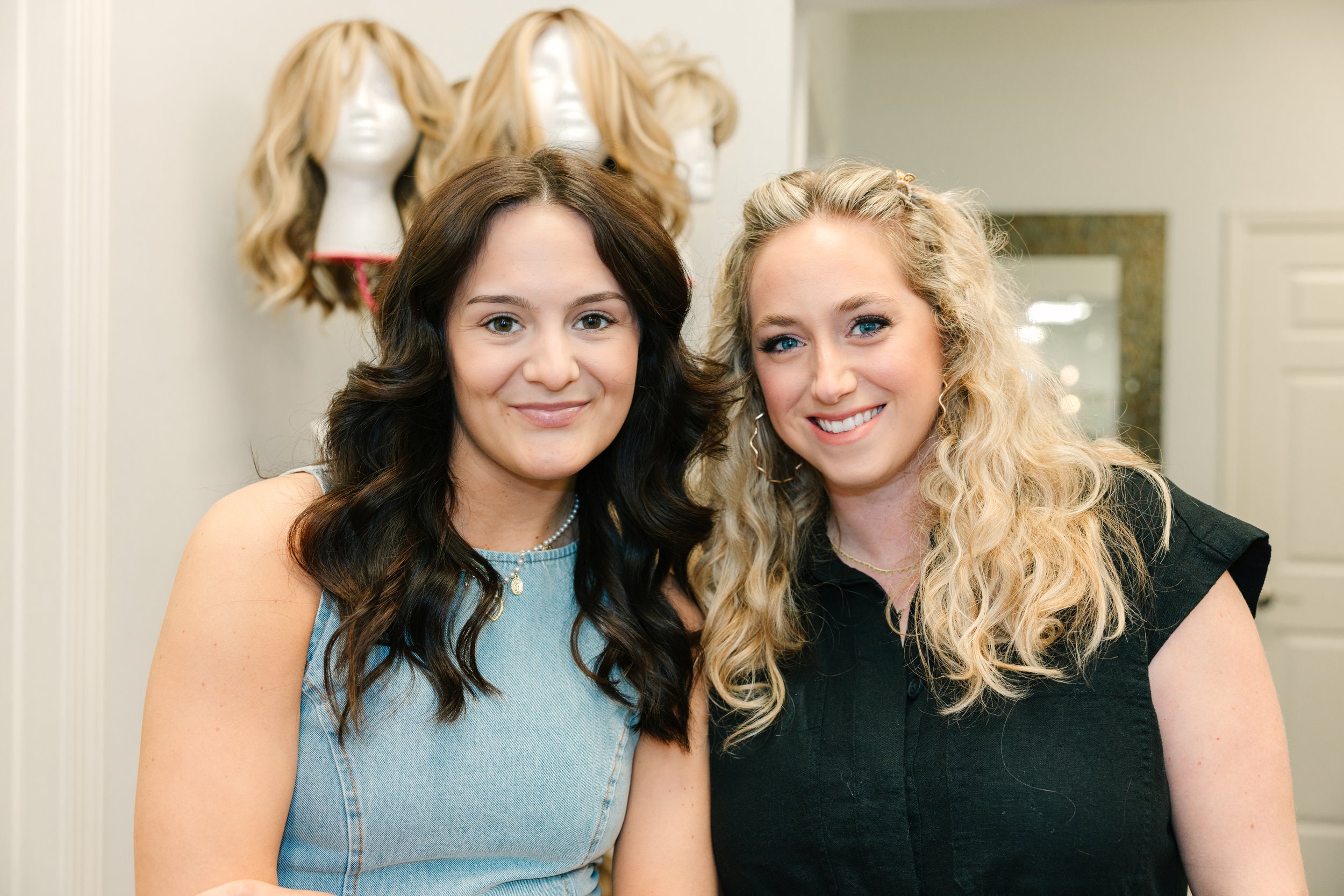 Two women smiling in front of mannequin heads with blonde wigs in a salon or wig shop.