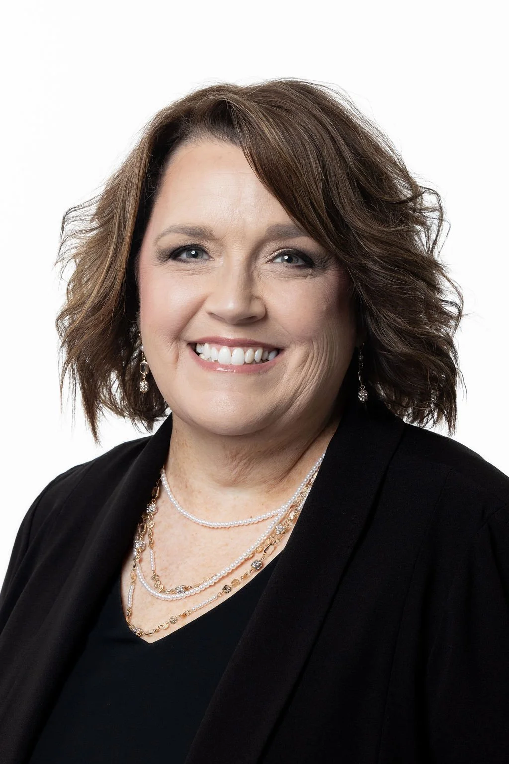 A portrait of a smiling woman with shoulder-length brown hair, wearing earrings and layered necklaces, dressed in a black top and black blazer against a white background.