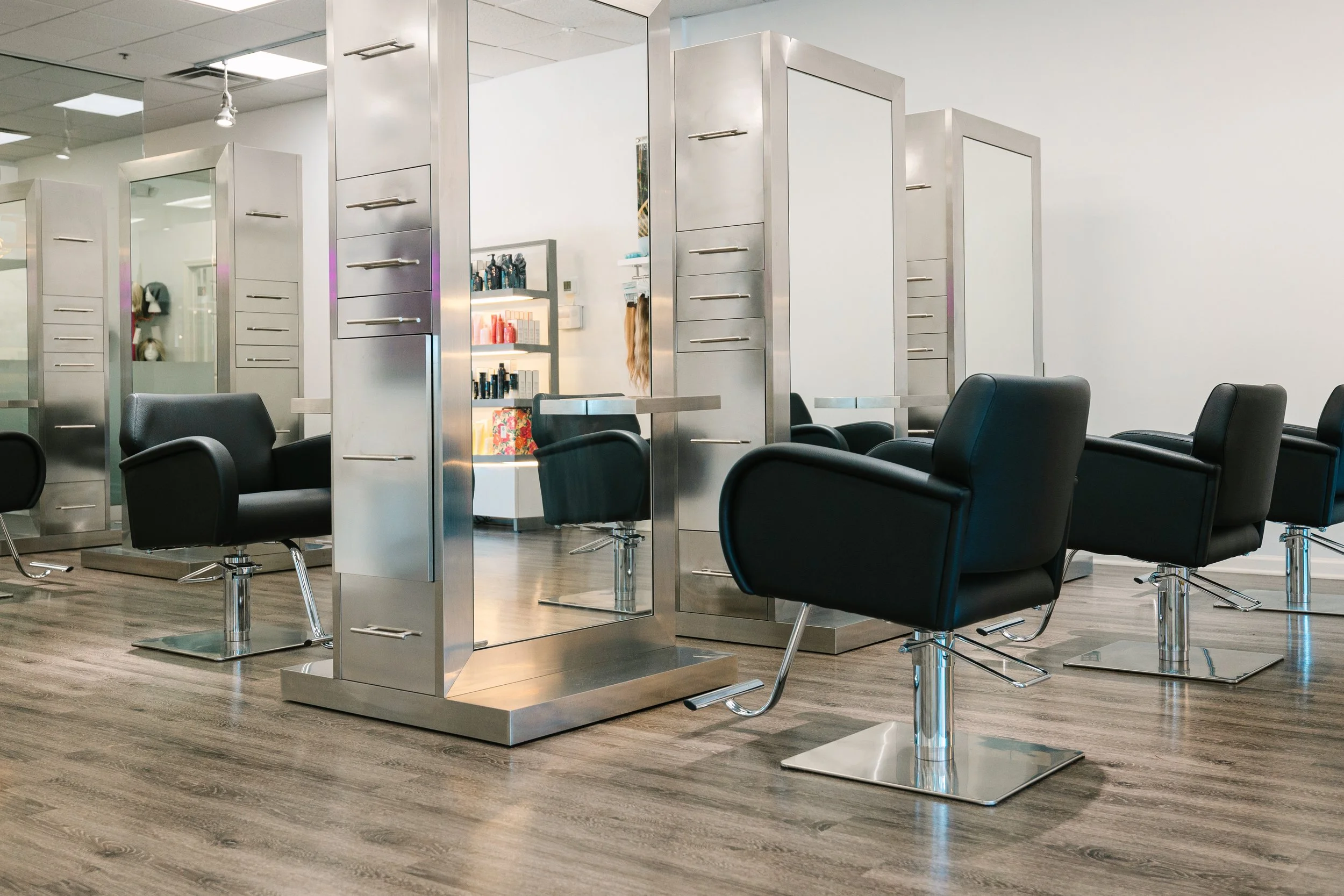 Jair salon with black salon chairs and mirrored stations, wood flooring, shelves with hair products in the background.