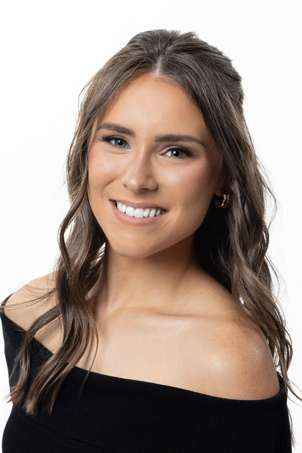 Close-up of a young woman with shoulder-length wavy brown hair, smiling, wearing a black off-shoulder top, and gold hoop earrings, against a white background.