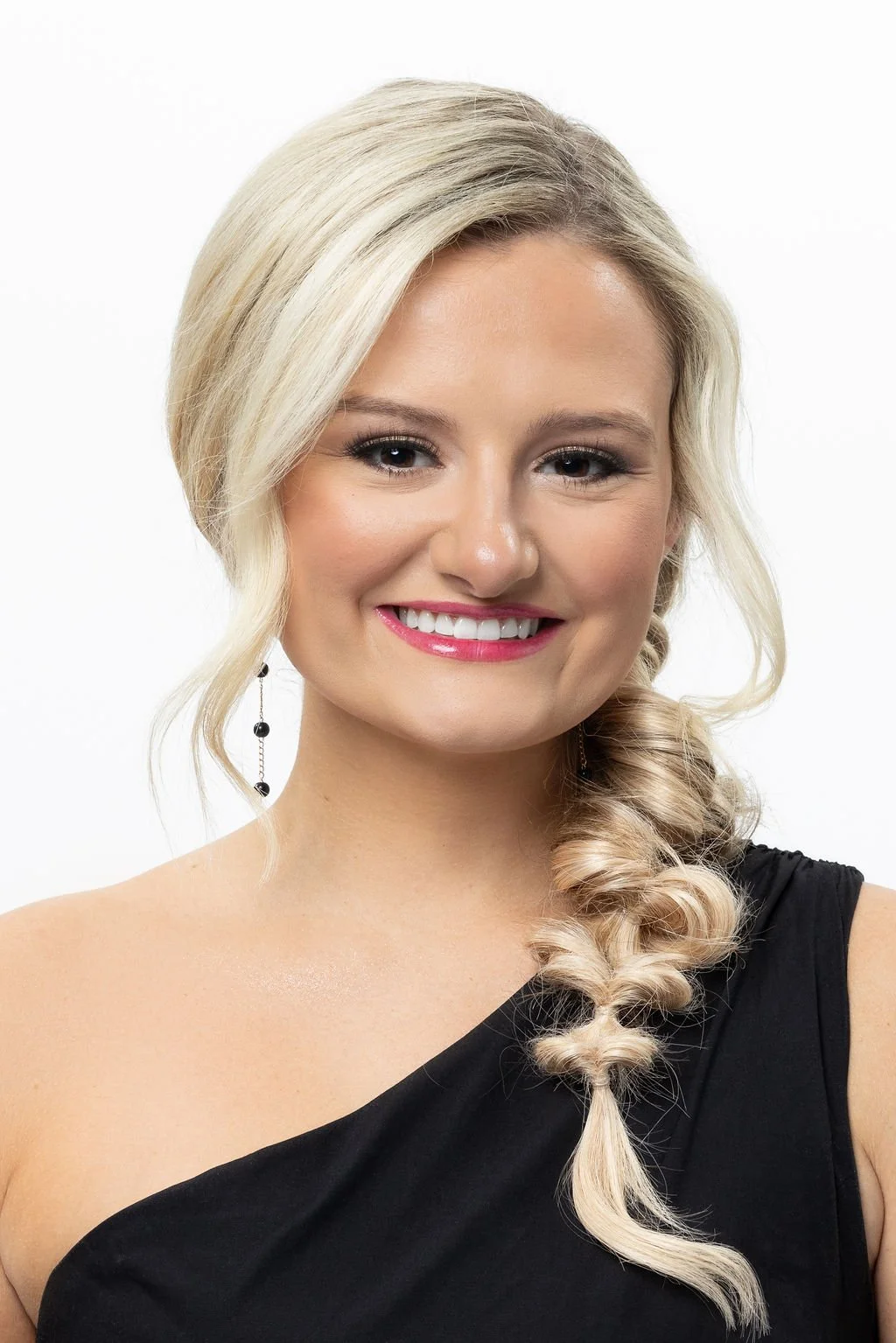 A young woman with blonde hair styled in loose curls, wearing a black sleeveless dress and dangling earrings, smiling against a plain white background.
