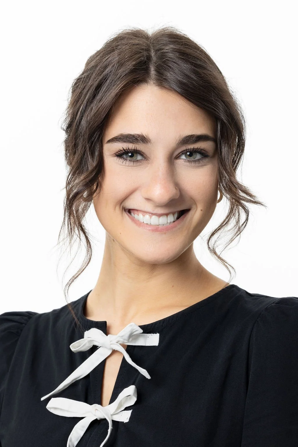 Smiling young woman with dark brown hair styled in loose waves, wearing a black top with white ribbon ties at the neckline.