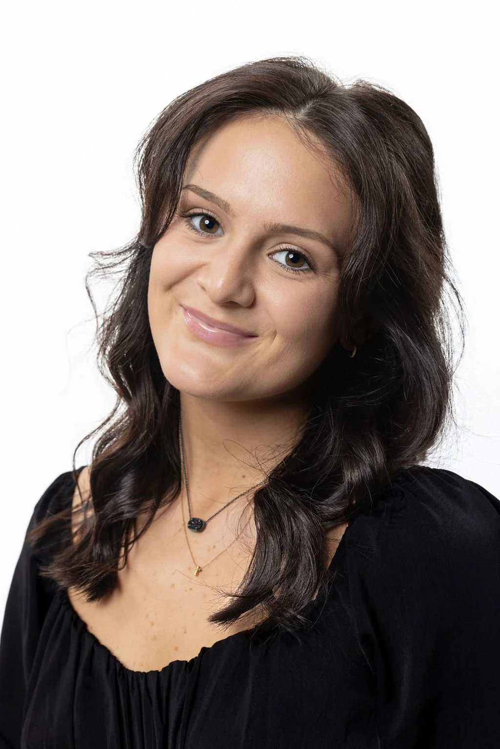 A smiling woman with medium-length dark brown hair and light eyes, wearing a black top and layered necklaces, posed against a white background.