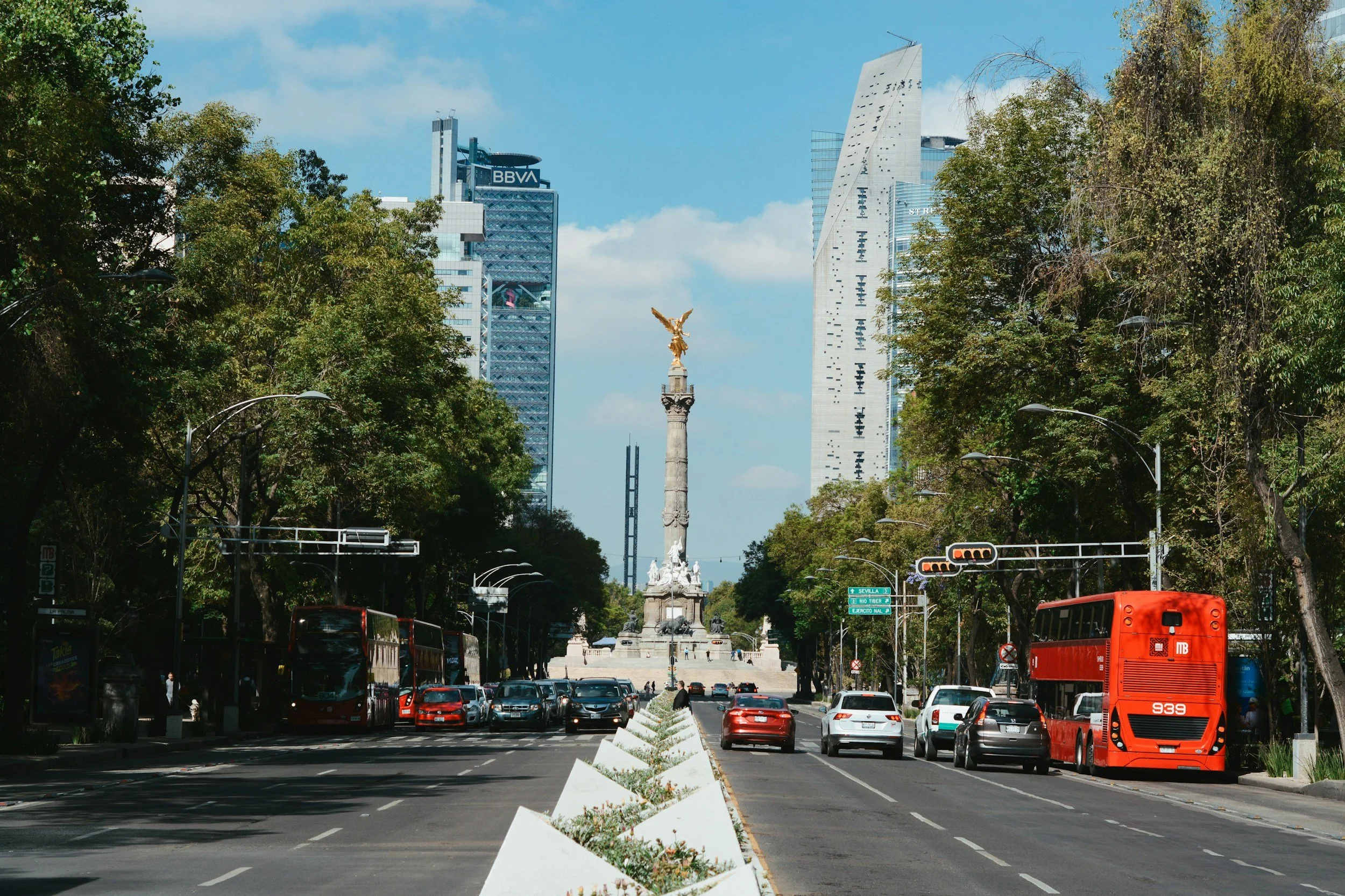 Street view of Mexico City with Monument to the Revolution in the background, skyscrapers, trees, red buses, and cars.