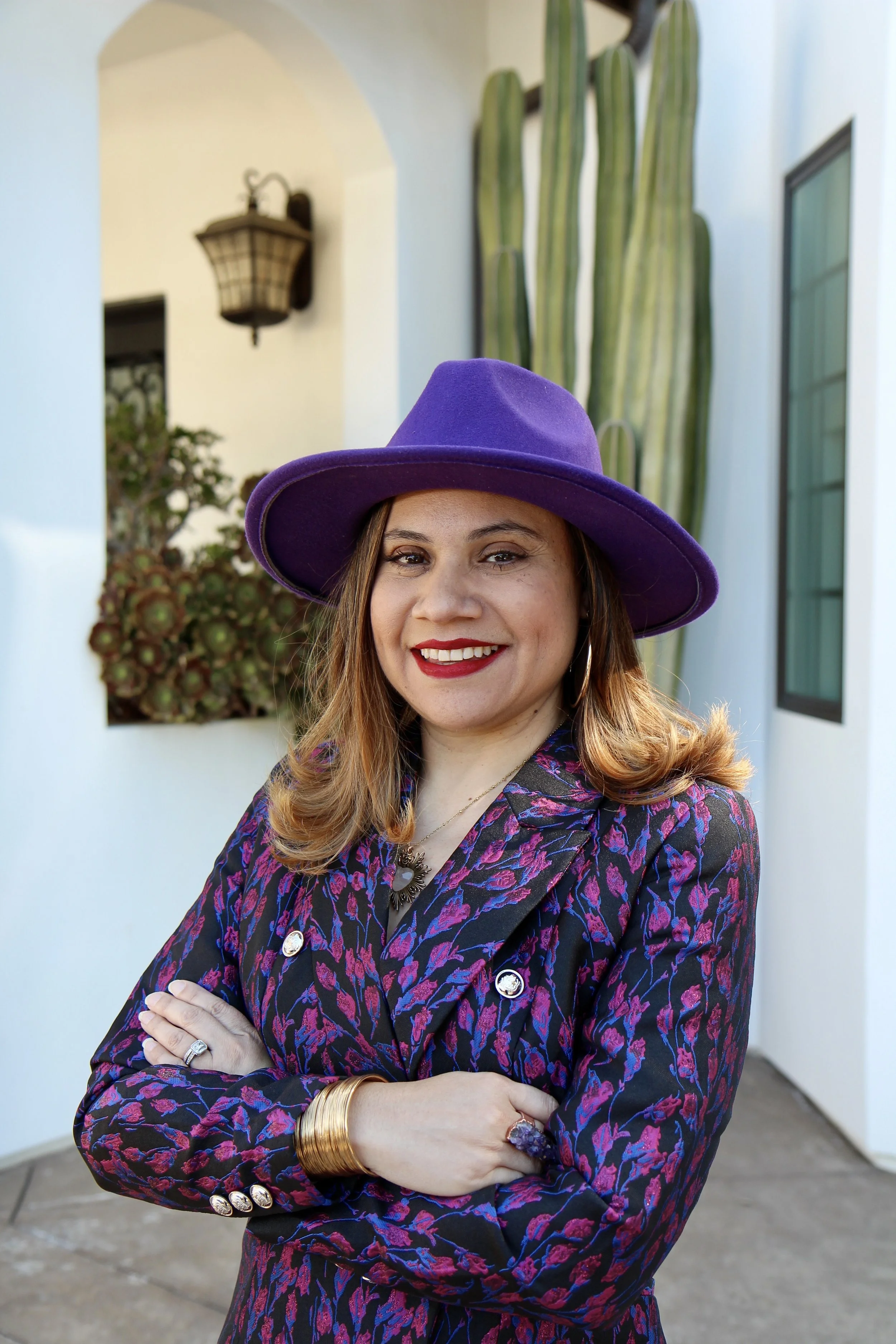 A woman wearing a purple hat and a patterned blazer stands outdoors with her arms crossed, smiling at the camera.