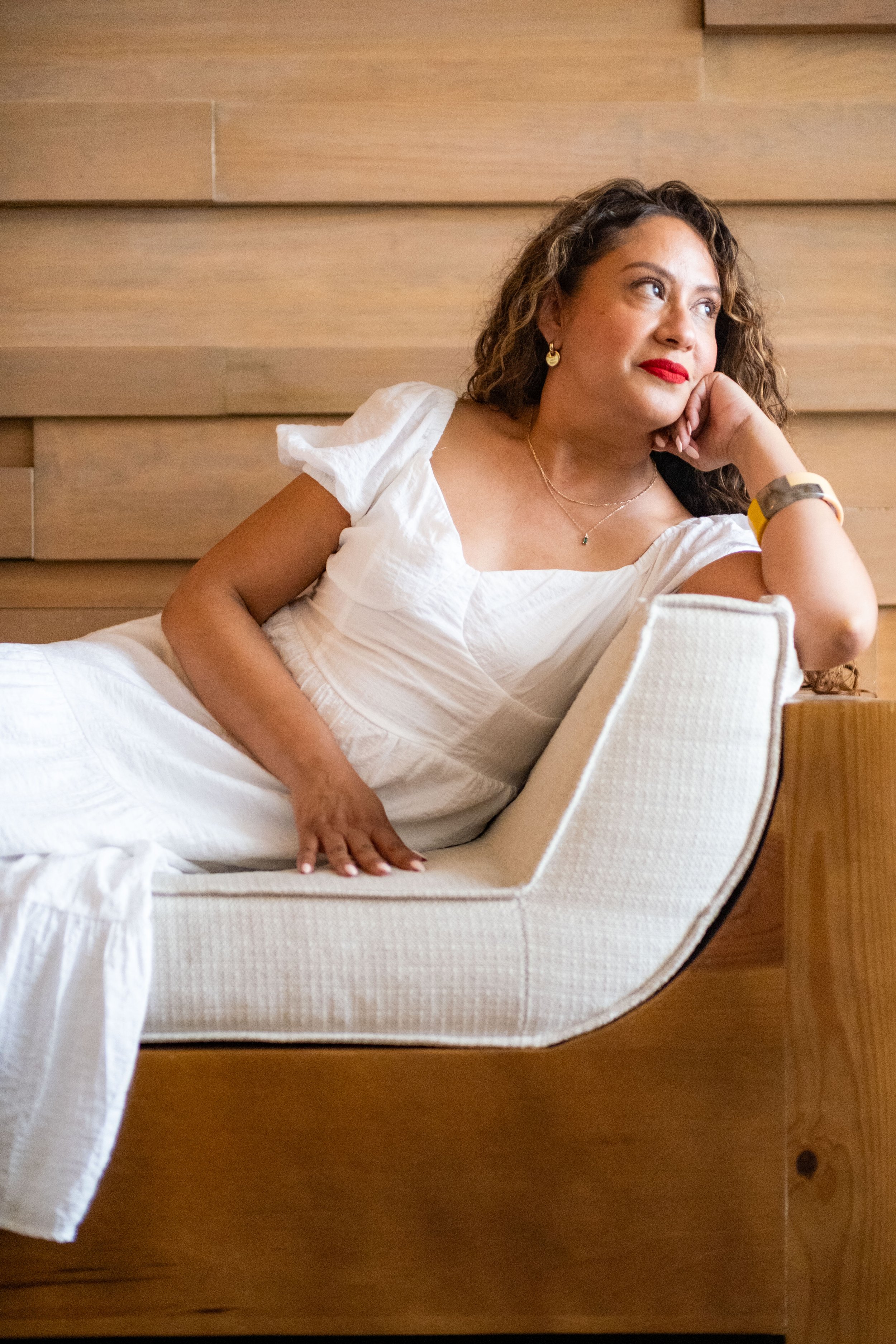 A woman with curly hair lying on a bed, resting her head on her hand, looking thoughtfully to the side. She is wearing a white dress, gold earrings, a necklace, and a watch, with a wooden-paneled wall in the background.