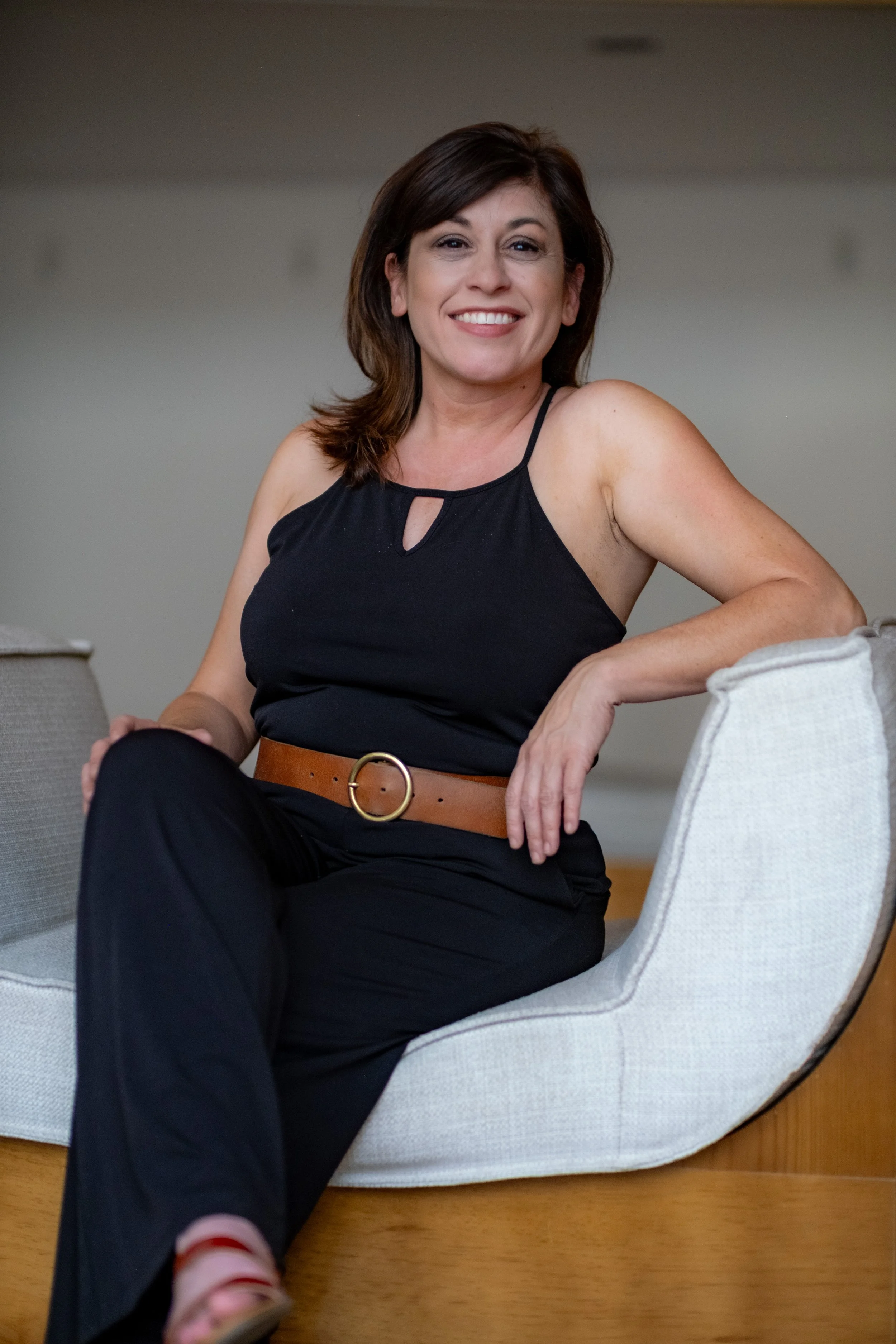 A woman with shoulder-length brown hair smiling while sitting on a light-colored sofa with her arm resting on the back.