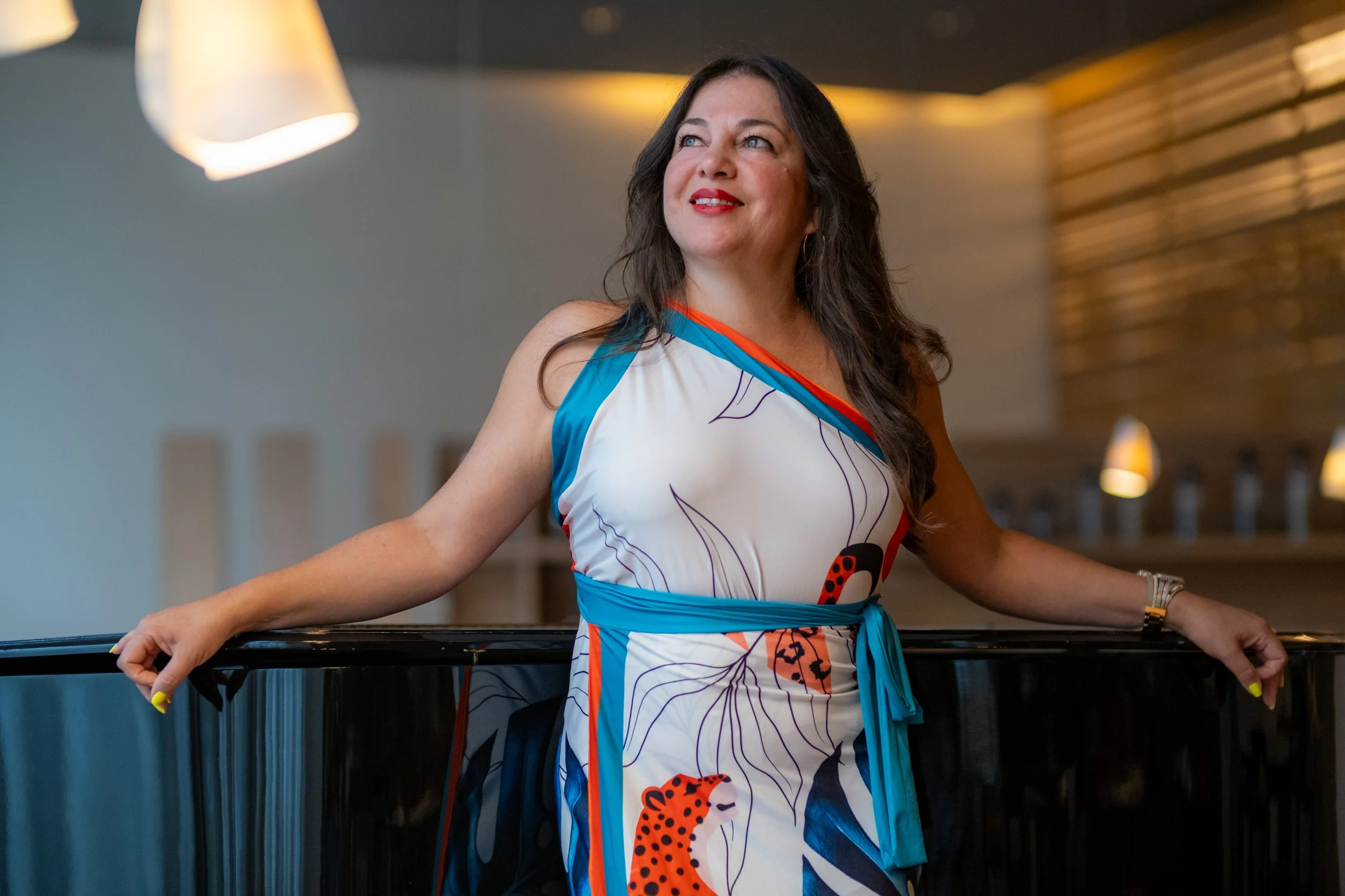 A woman with long dark hair wearing a colorful, patterned dress with jaguar and leaf designs, leaning on a black piano with her arms resting on it, in a modern, softly lit interior.