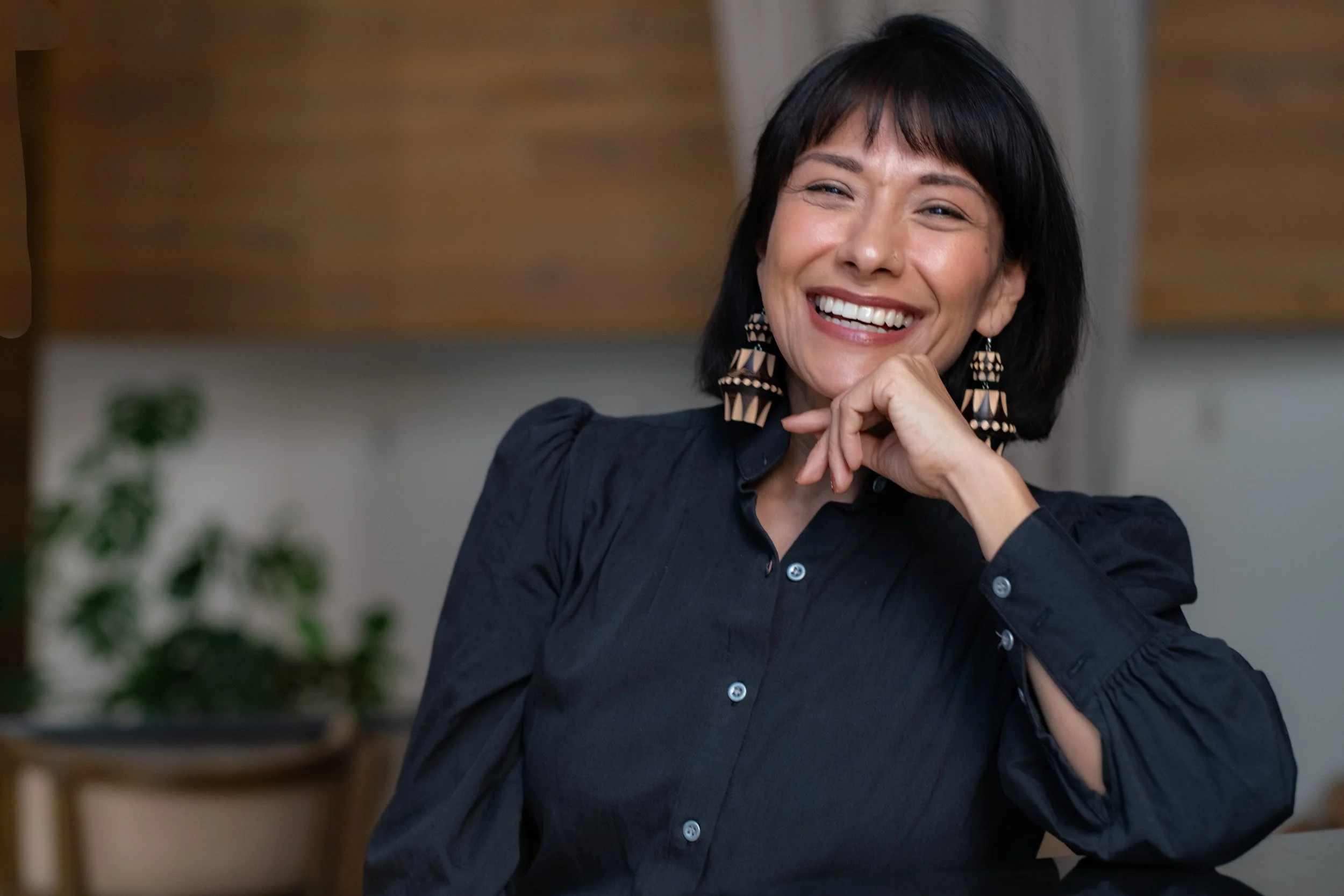 A woman with short black hair and a big smile, wearing a dark button-up shirt and large patterned earrings, sitting indoors in a modern setting.