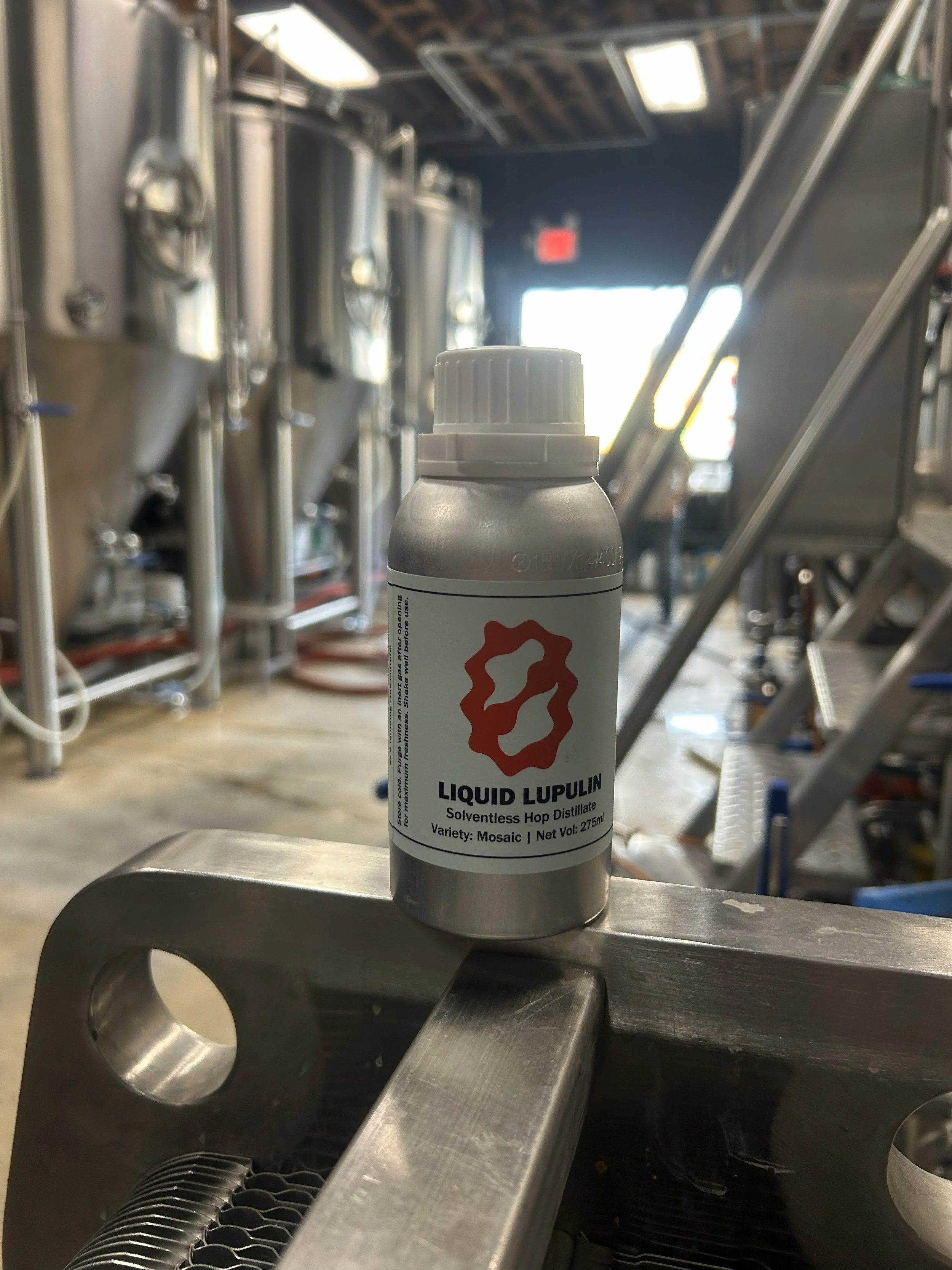 A bottle labeled 'Liquid Lupulin, Solventless Hop Distillate, Mosaic' resting on a metal surface in a brewery or distillery setting.