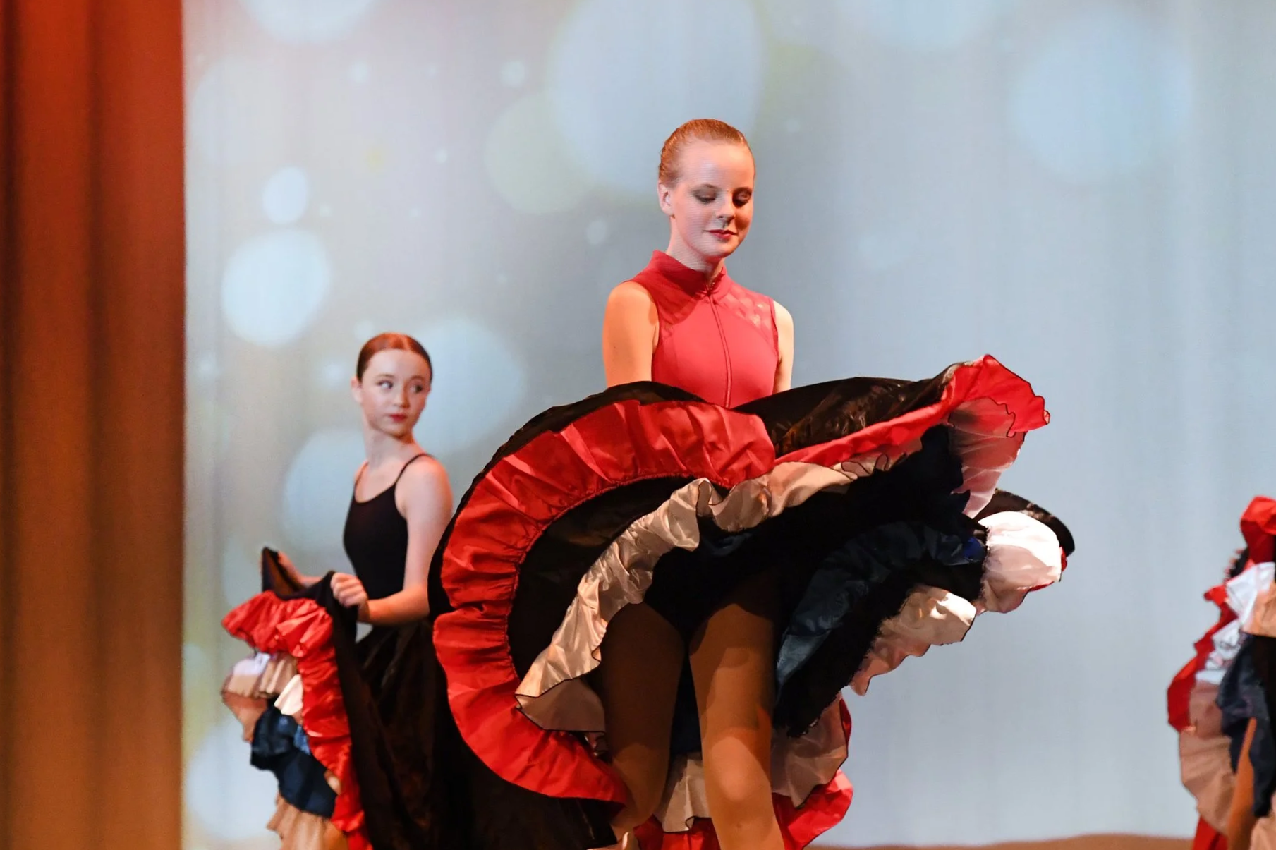 Group of young women performing a dance on stage, dressed in colorful skirts and dance shoes, with a projected background of bokeh circles.