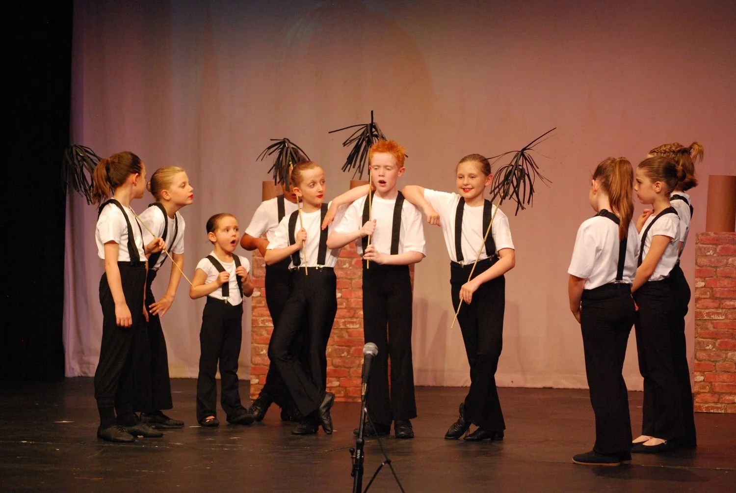 Children performing on stage, dressed in black suspenders and white shirts, with some holding sticks with paper palm leaves, during a theatrical play.