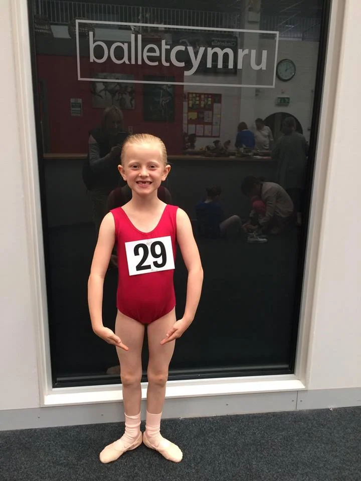 Young girl in a red gymnastics leotard with a number 29 badge, standing in front of a glass window with the words "balletcymru."