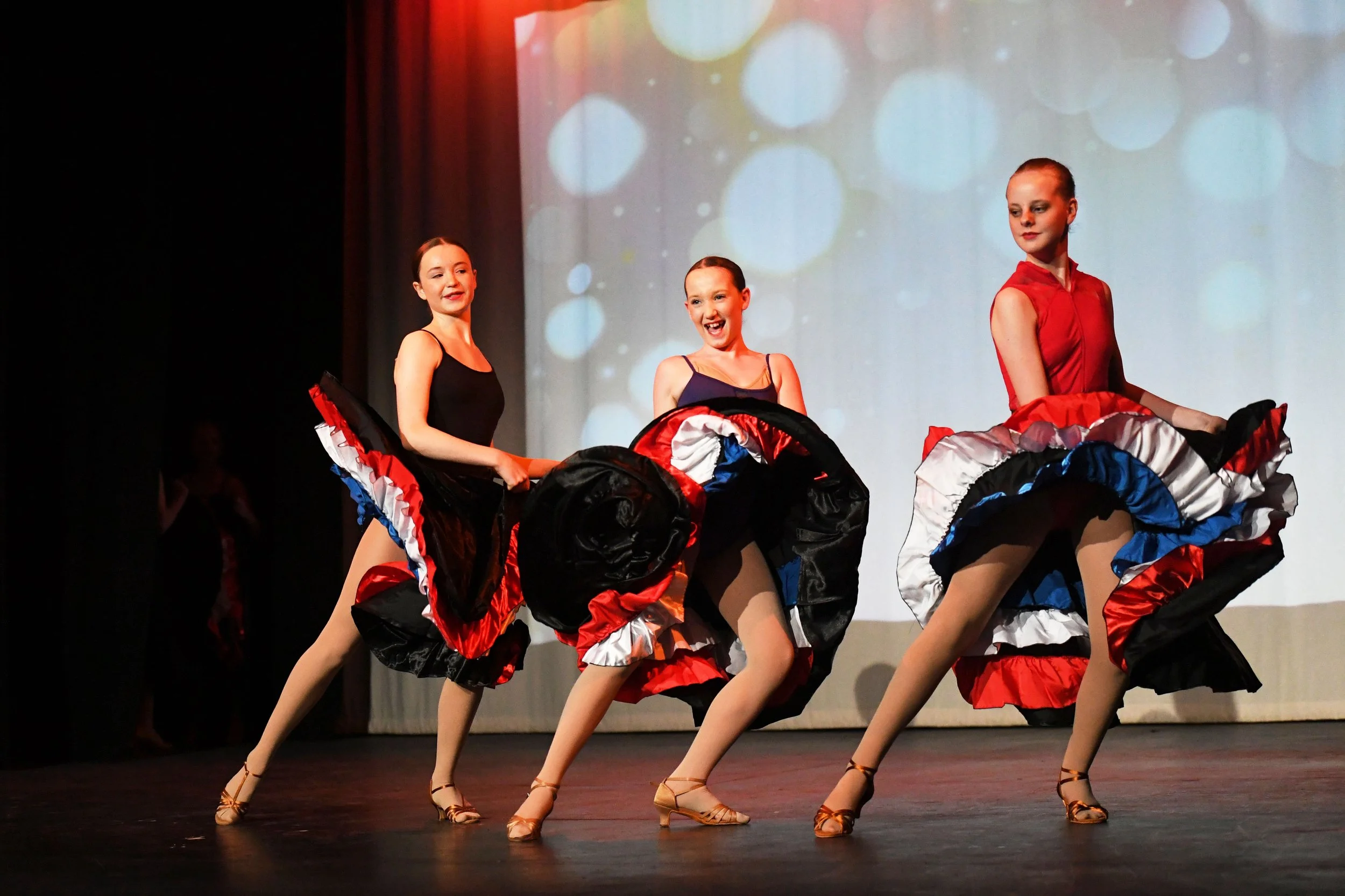 Group of young women performing a dance on stage, dressed in colorful skirts and dance shoes, with a projected background of bokeh circles.