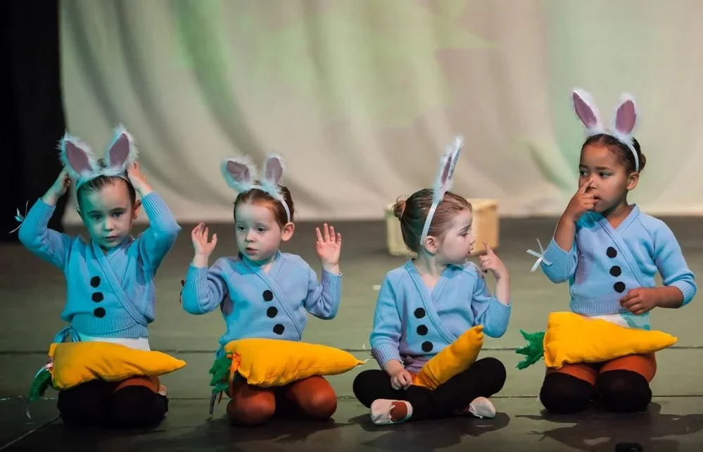 Four young girls dressed as rabbits sitting on stage, holding plush carrot props, preparing for a performance.