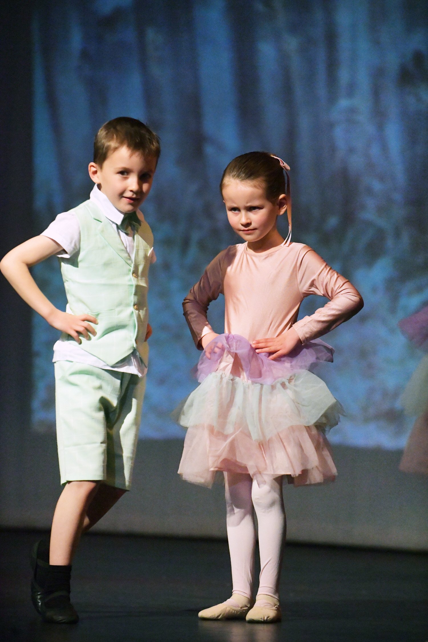 Two children standing on stage, a boy on the left and a girl on the right, both dressed in costumes with a blue curtain backdrop.