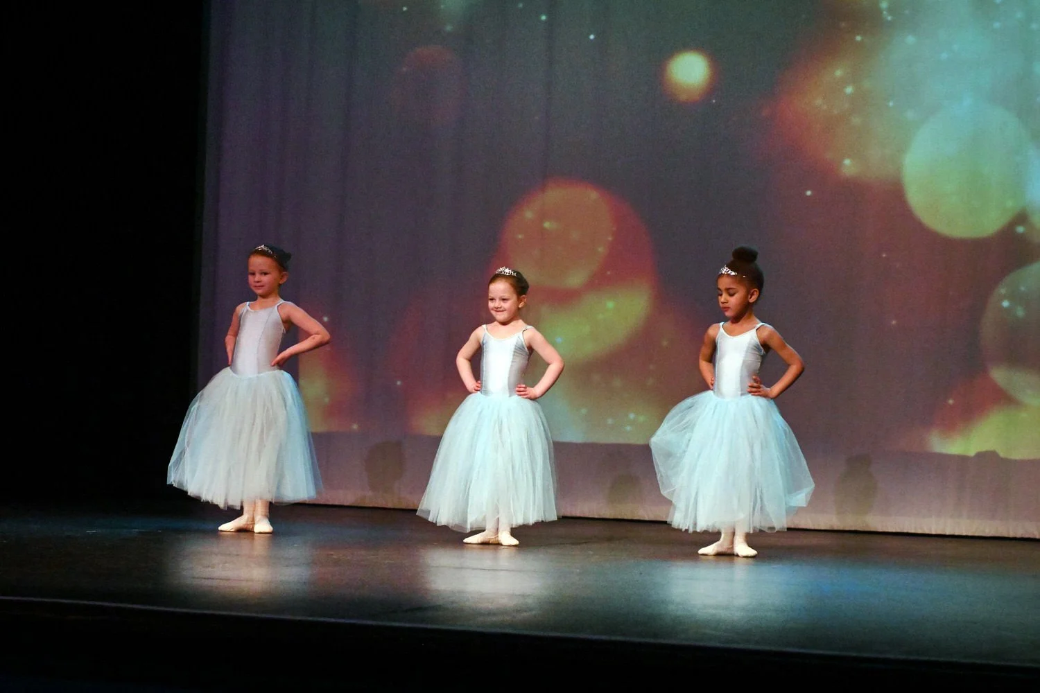 Three young girls in ballet costumes performing on a stage with a colorful, abstract background.