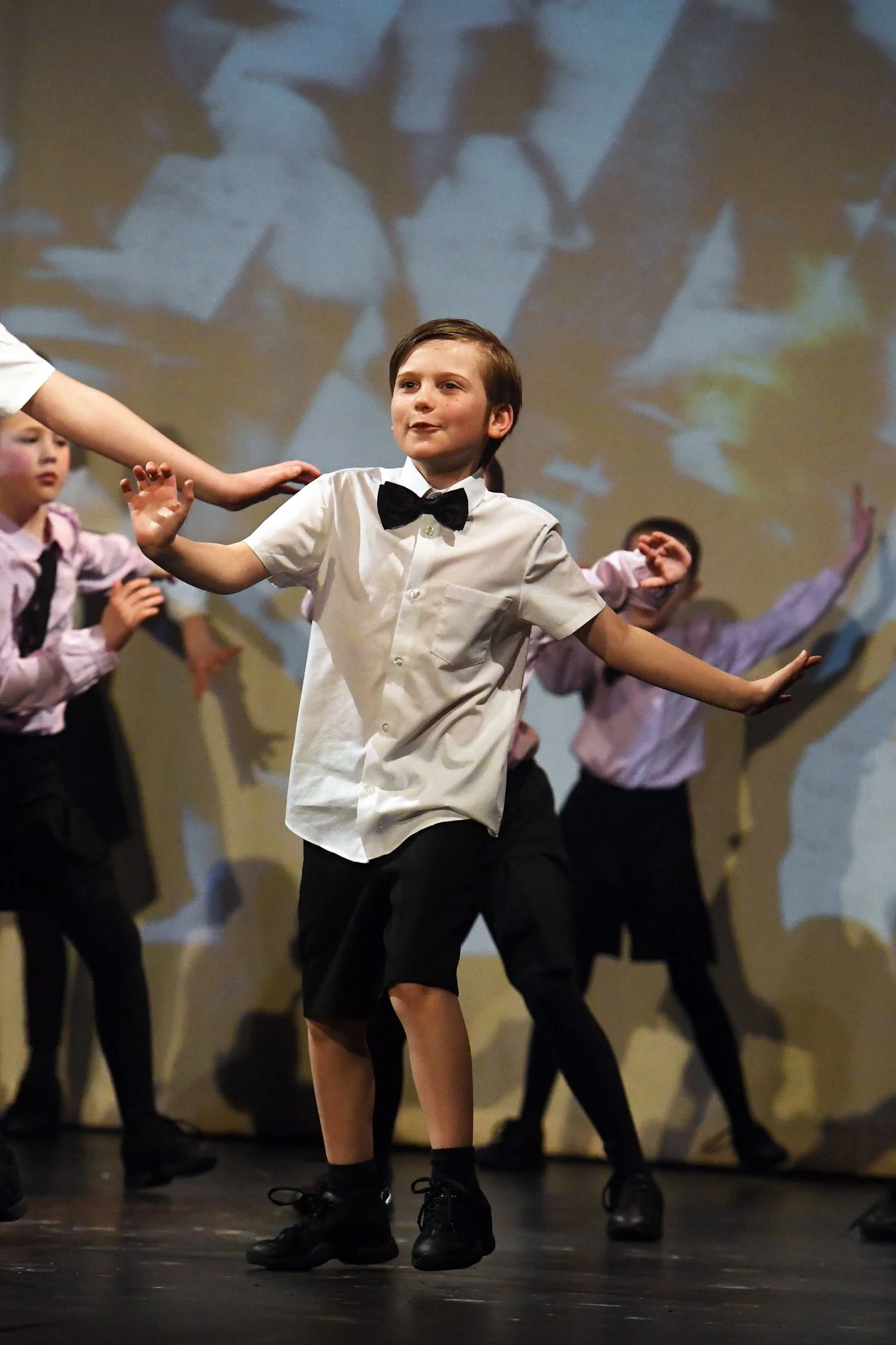 Children performing a dance on stage, with one boy in focus wearing a white shirt, black bow tie, and black shorts, leading the group, while others in the background wear similar outfits and move in coordination.