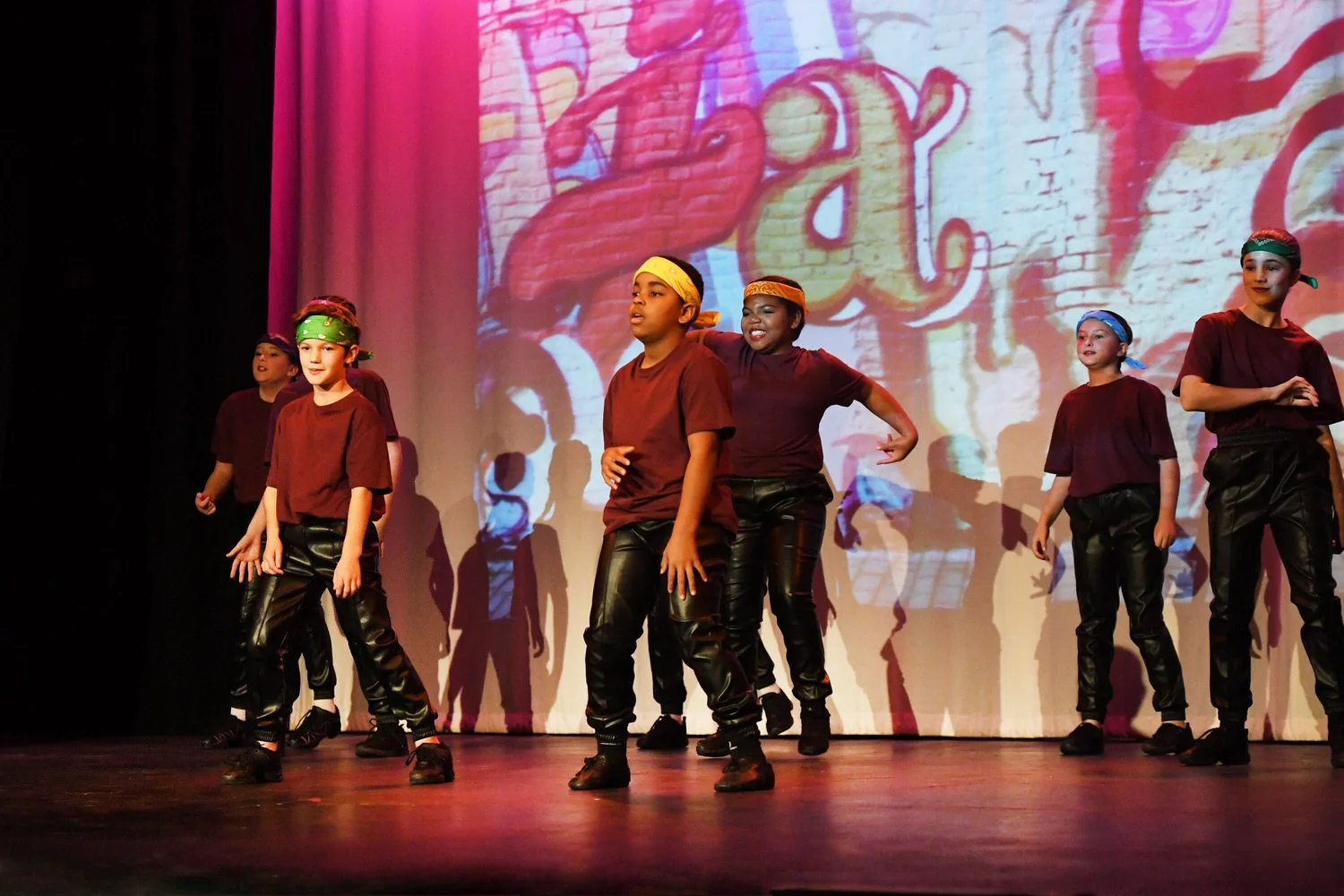 Group of children performing a dance on stage during a school play or recital, with a colorful graffiti-style background projected on the screen behind them.