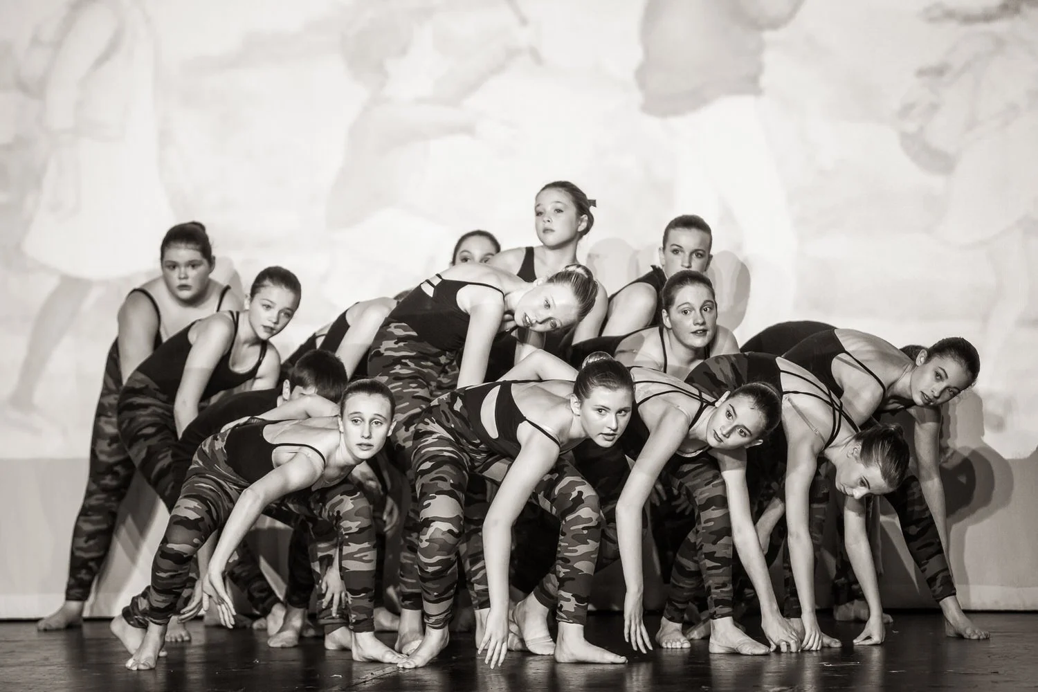 Black and white photo of a group of young female dancers performing on stage in matching camouflage-patterned pants and black tops, arranged in a formation.