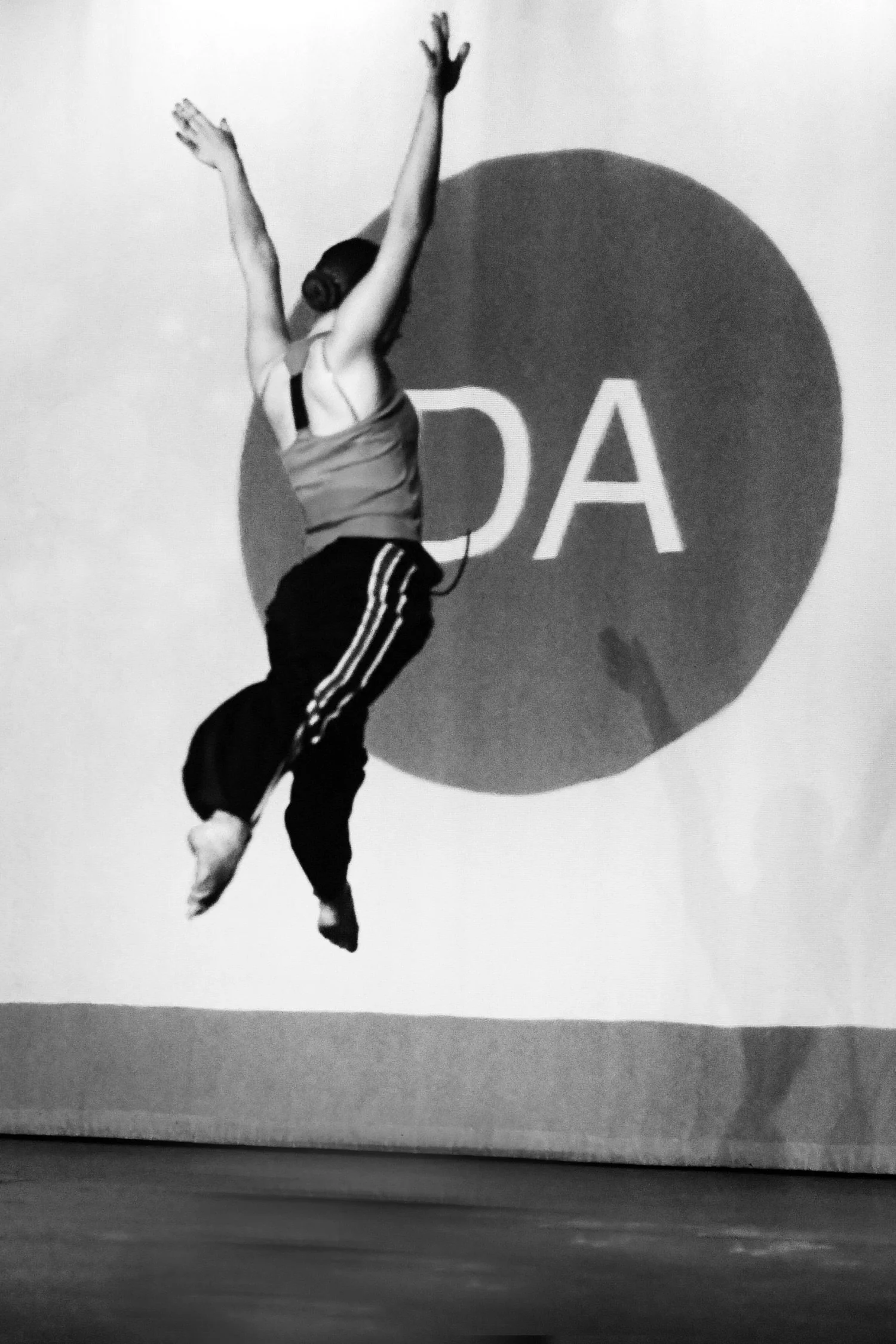 Black and white photo of a woman jumping in front of a wall with large text that reads 'DADA', shadowing her silhouette.