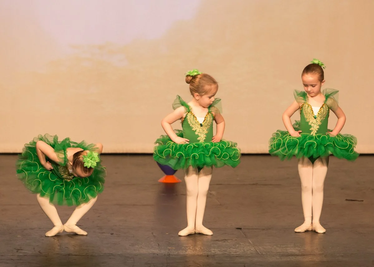 Three young girls in green ballet costumes performing on stage, with the girl on the left bending over and the other two standing with hands on hips.
