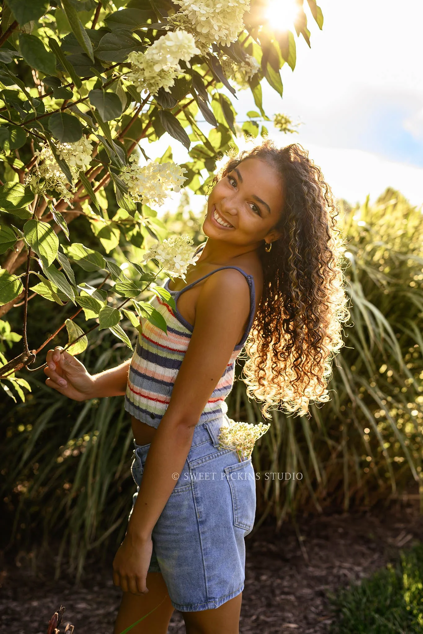 Madeline | Indiana Floral Senior Girl Photography posing by flowers for Speedway High School Class of 2027 wearing striped tank top and denim shorts by Sweet Pickins Studio senior photographer Amanda Wilson ©