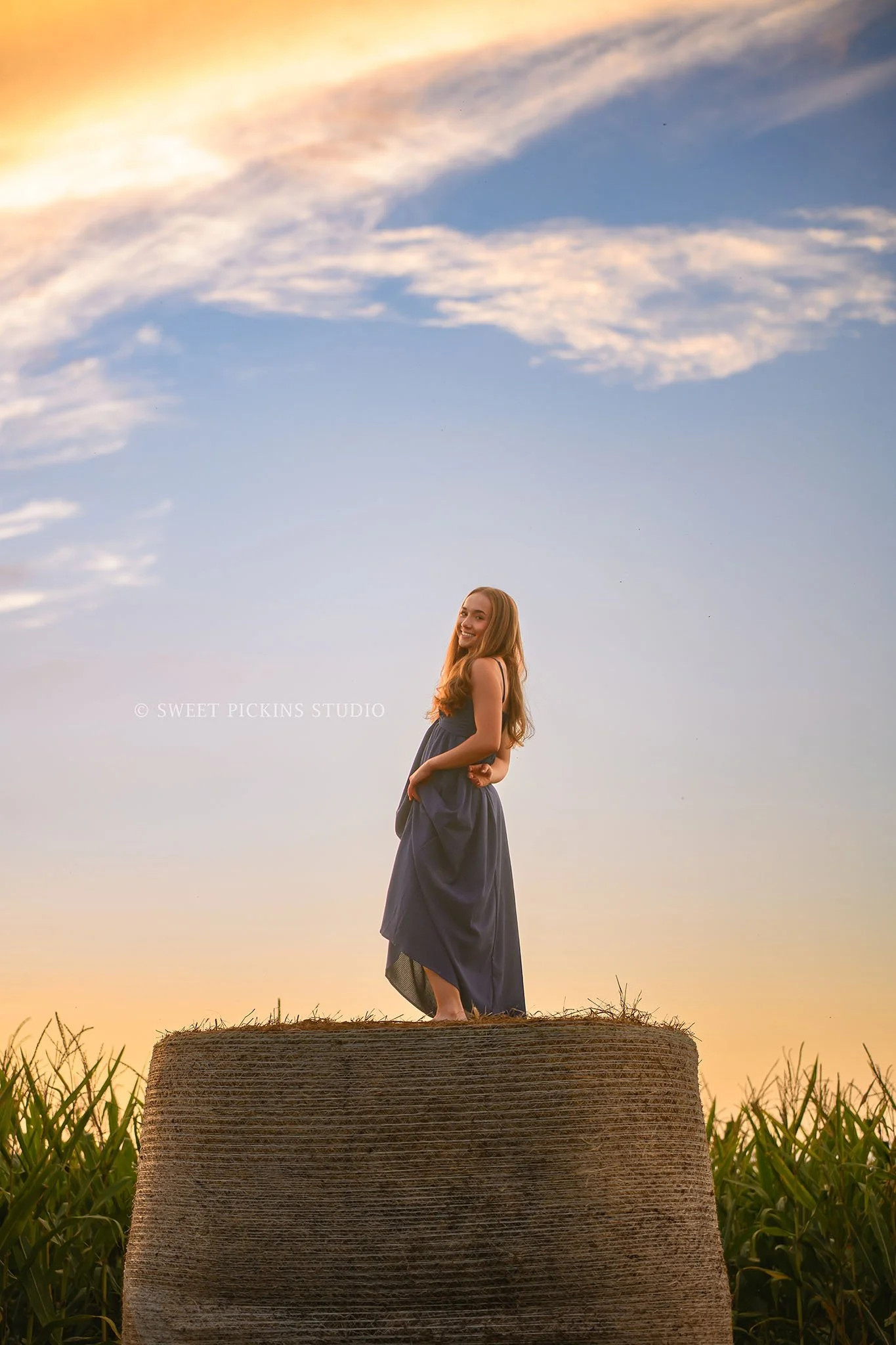 Natalie’s Lowell, Indiana Farm Senior Photography posing on hay bales by corn in blue dress at sunset  by Sweet Pickins Studio senior photographer Amanda Wilson ©