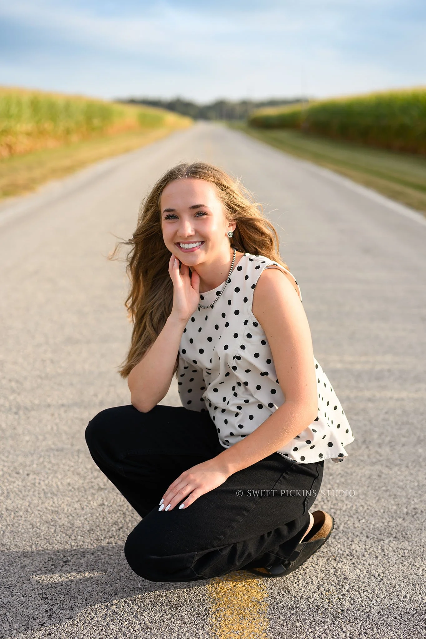 Natalie’s Lowell, Indiana Senior Girl Photography for class of 2027 posing in the middle of country road wearing black polka dot top by Sweet Pickins Studio senior photographer Amanda Wilson ©